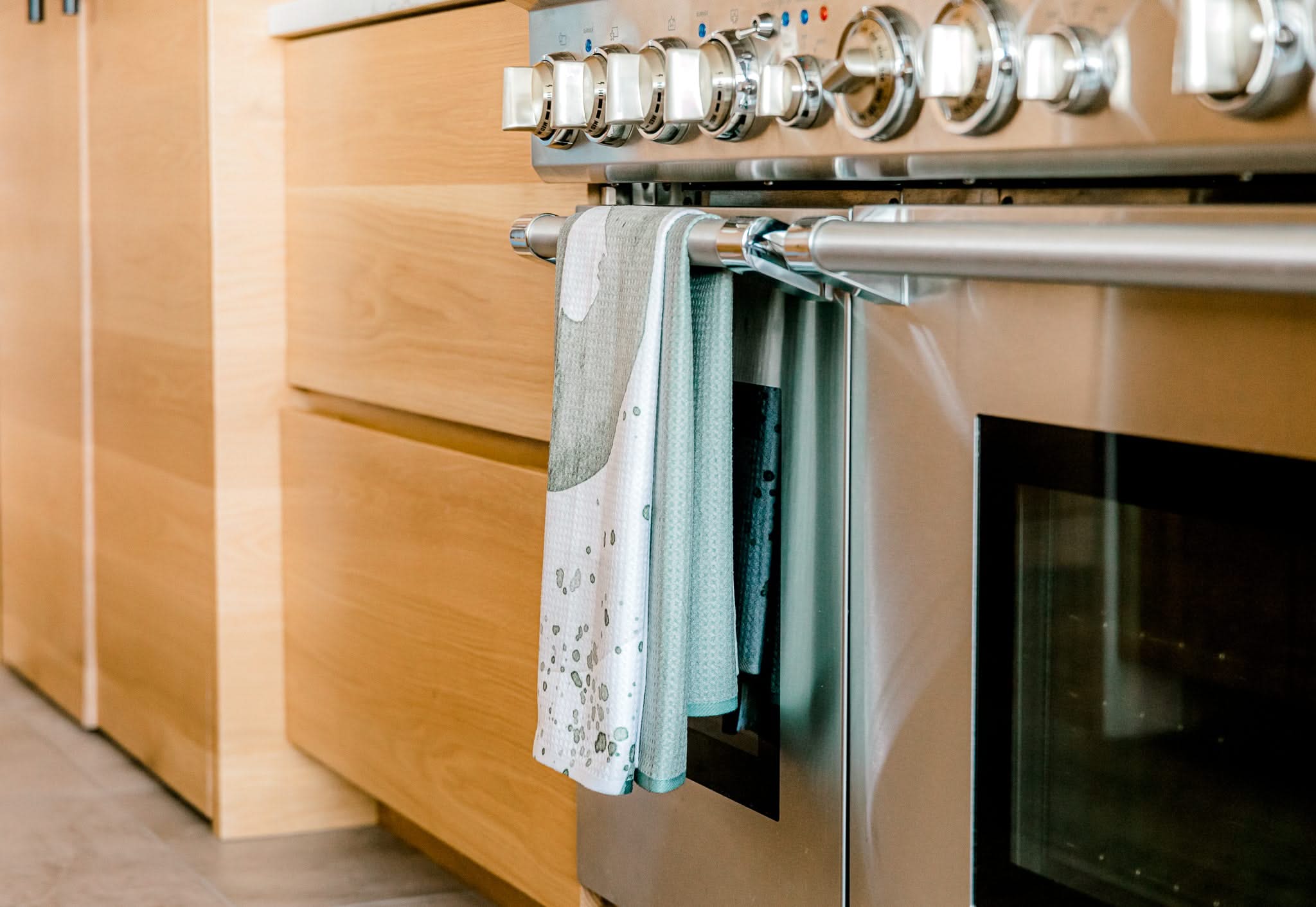 A stainless steel oven with a glass door and control knobs is shown. Two Kailee Wright Hunter Abstract Tea Towels, crafted from Geoweave fabric, hang from the oven handle. Light wood cabinets appear beside the oven.