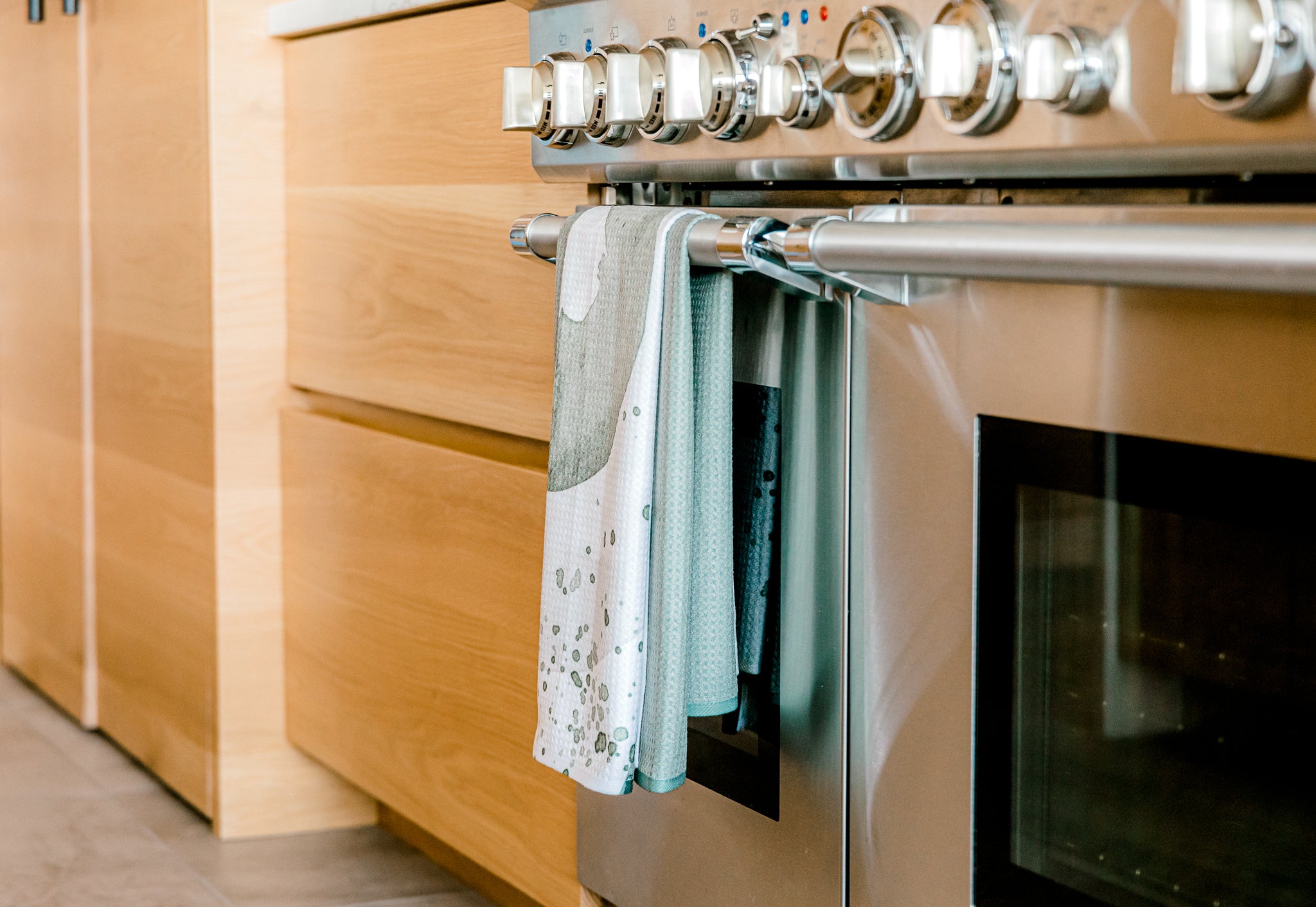 A Kailee Wright Hunter Green super-absorbent dishcloth hangs from the handle of a stainless steel oven with a glass door, beside light wood cabinets in a modern kitchen.
