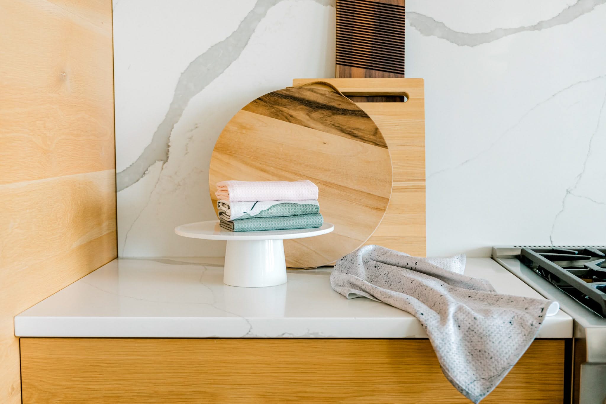 A modern kitchen counter displays a Kailee Wright Hudson Grey towel near the stovetop, with folded pastel Geoweave dish towels, a white cake stand, round and rectangular wooden cutting boards, and sleek marble and wood surfaces.