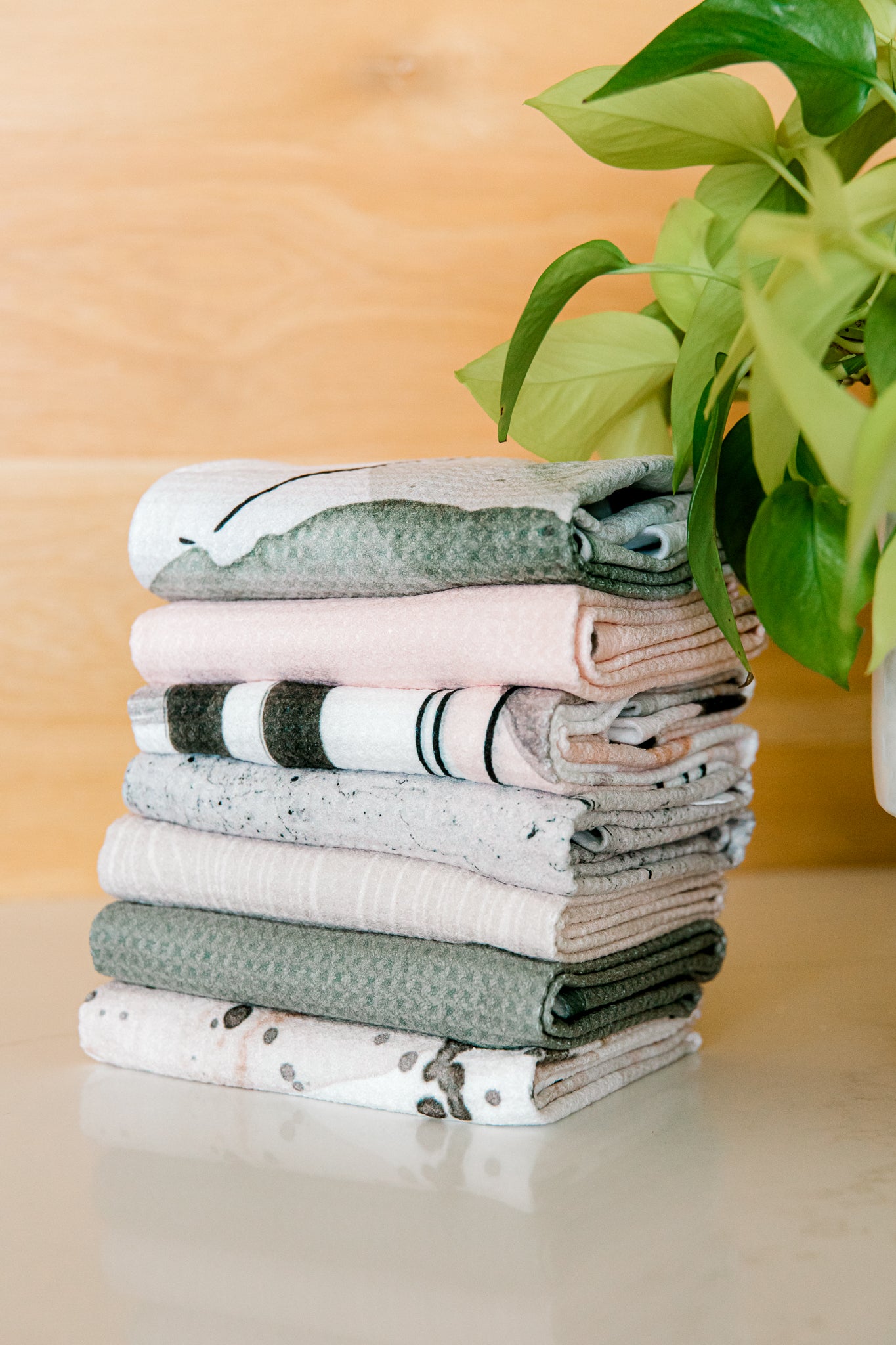 A neatly folded stack of Kailee Wrights Hunter Green towels made from super-absorbent Geoweave fabric sits on a white surface beside a green leafy potted plant, with a wooden background behind them.
