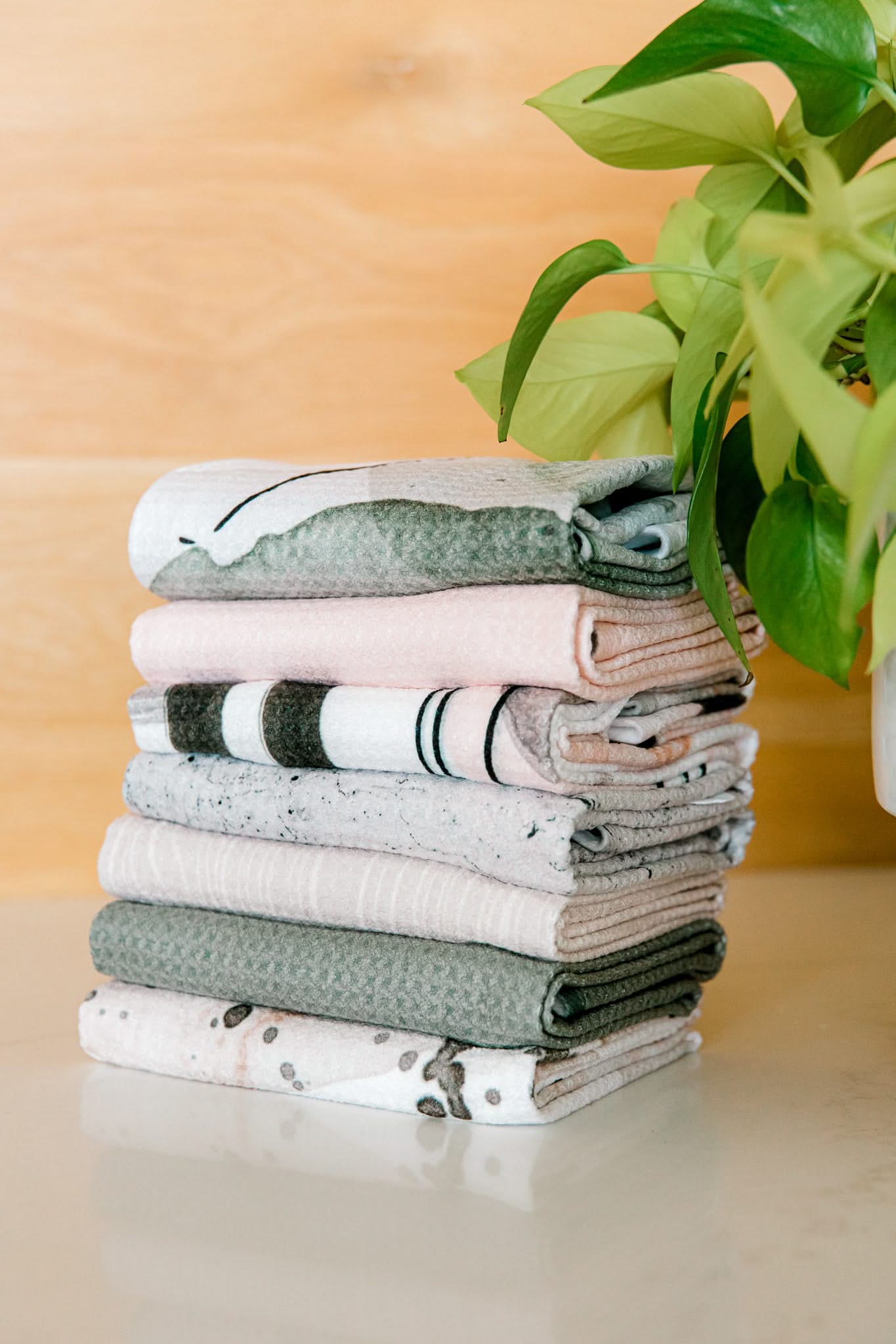 A neatly folded stack of Kailee Wrights Hunter Green towels made from super-absorbent Geoweave fabric sits on a white surface beside a green leafy potted plant, with a wooden background behind them.