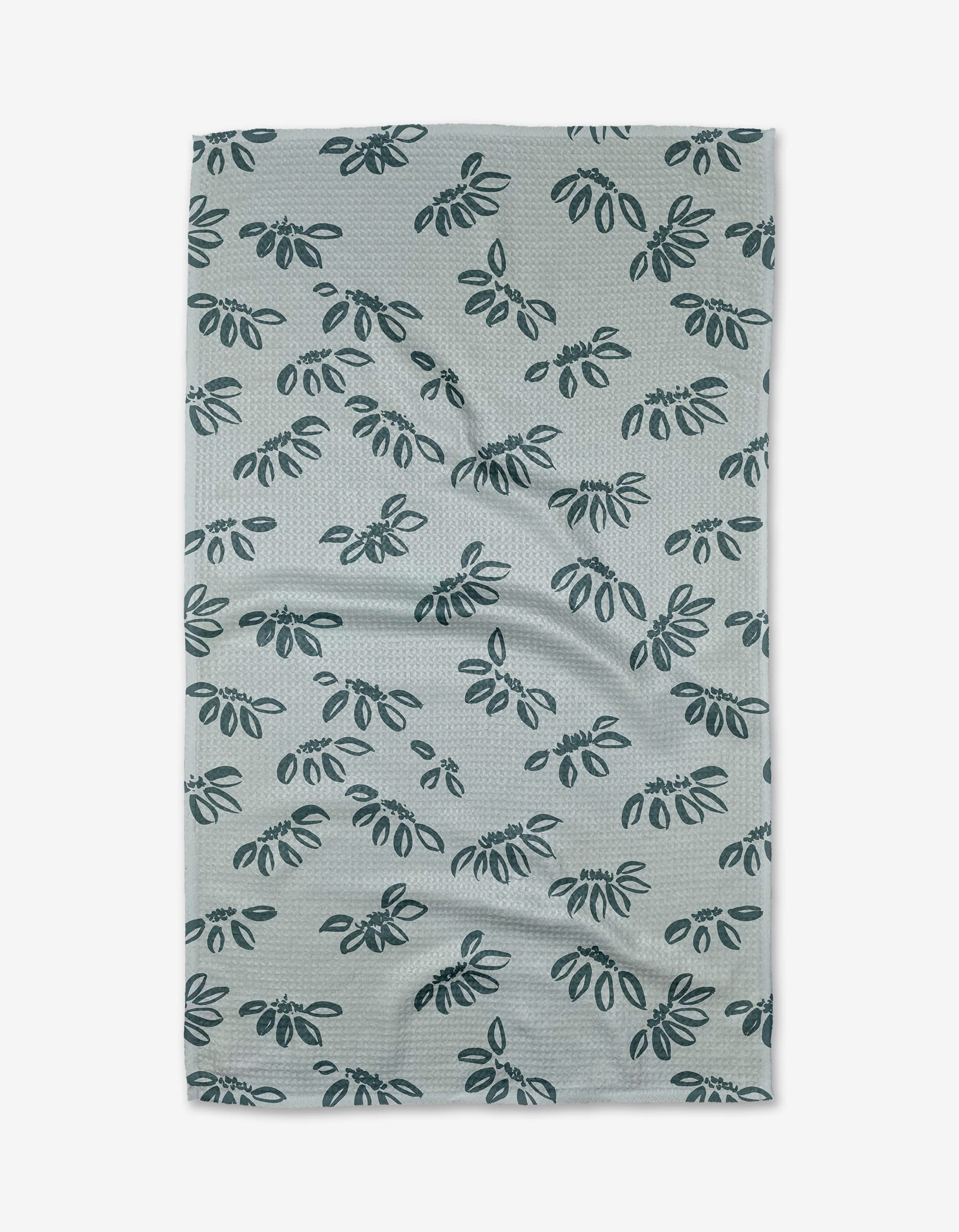 The Discovery Tea Towel by Toboggan Avenue is light gray and made from recycled Geoweave™ fabric, featuring a repeated dark blue leafy branch pattern. Shown flat with visible wrinkles.