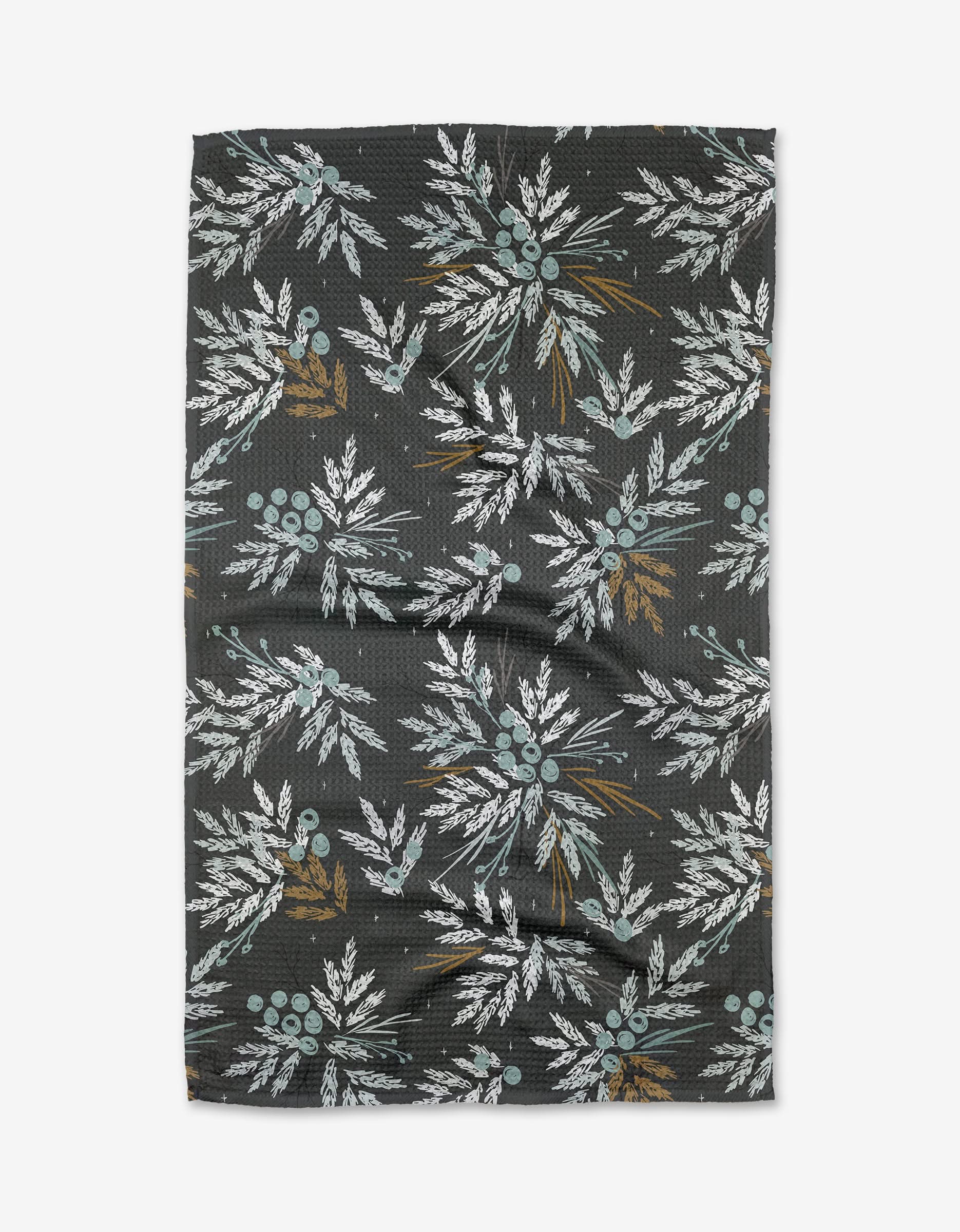 The “Frost” towel by Toboggan Avenue is dark-colored, quick-drying, and features a repeating pattern of white leafy branches with muted blue and brown berries on a subtly textured surface.