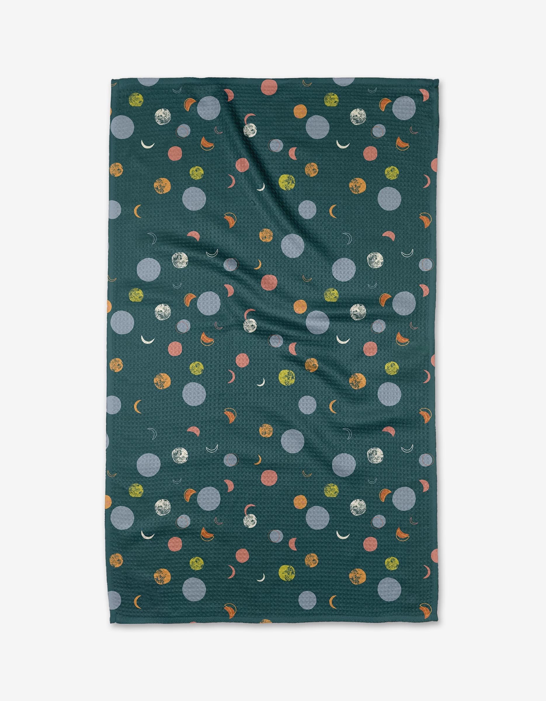 The Lumina kitchen towel by Jessica Swift is a dark green, rectangular towel with orange, yellow, blue, and white dots, crescent moons, and textured circles. Made with recycled materials; fabric has slight wrinkles. Photographed on a white background.