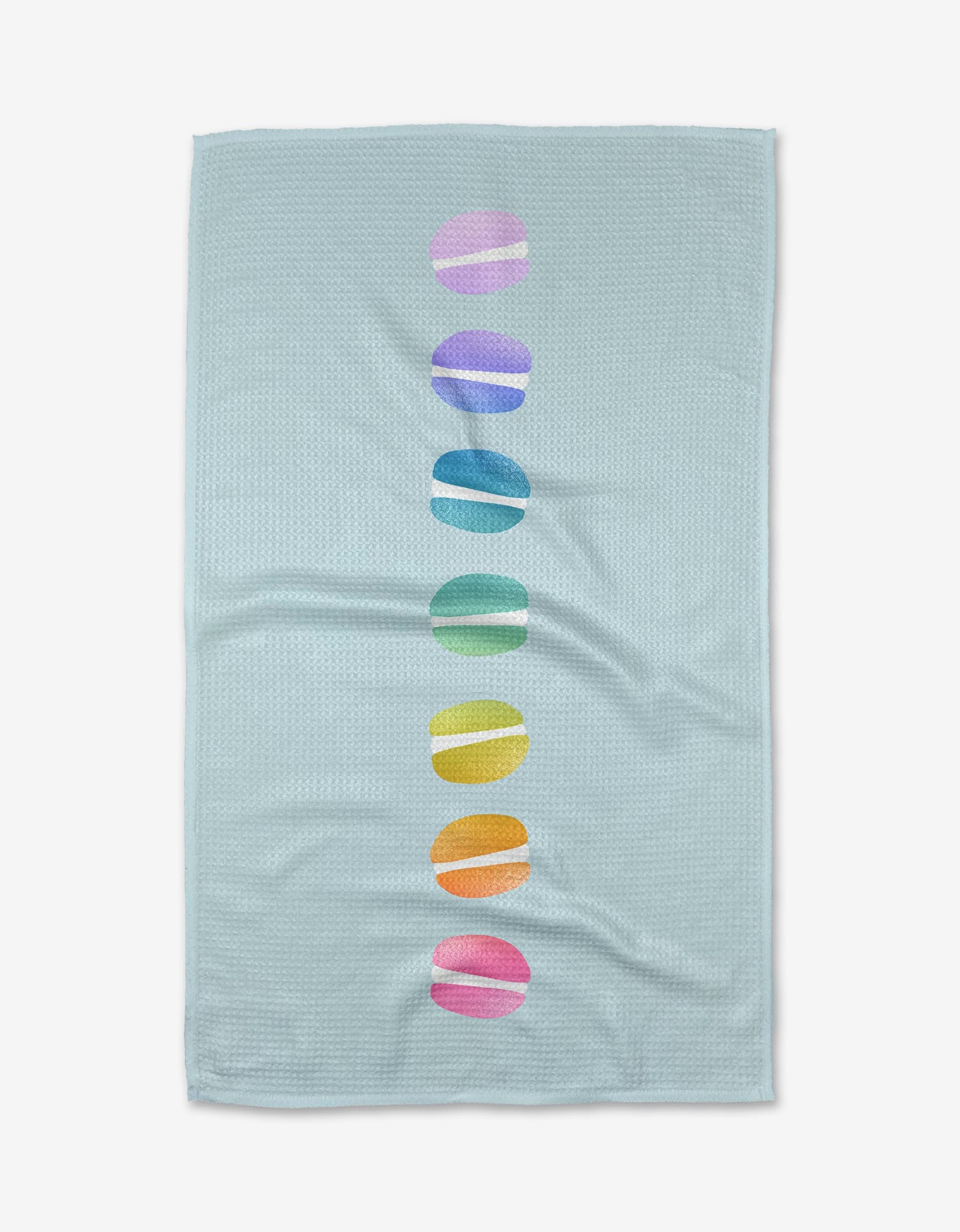 The Mac Love Kitchen Tea Towel by Noras Macarons features light blue Geoweave fabric with a centered vertical row of seven colorful, rainbow-hued macaron ovals.