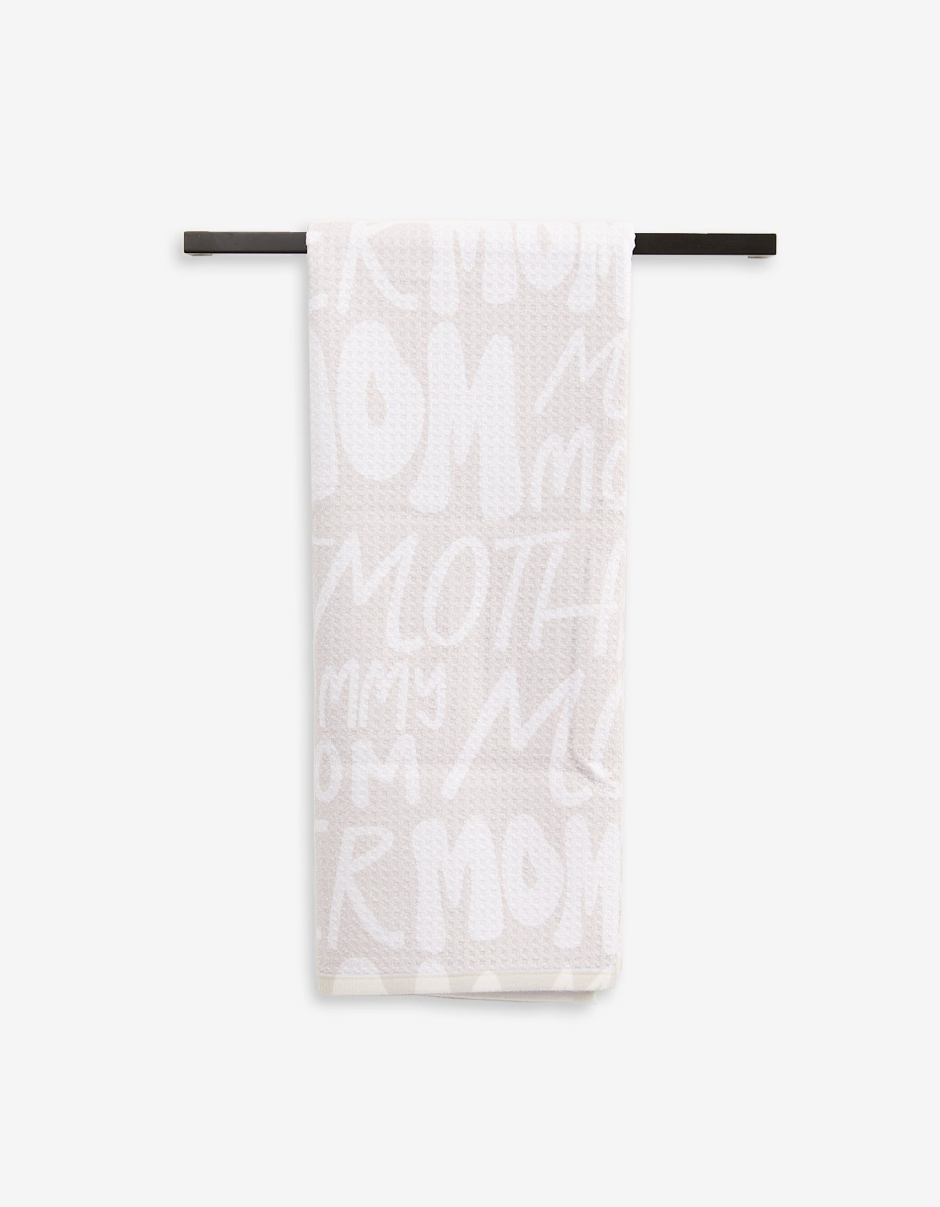 A light-colored, ultra-absorbent GEOMETRY Mom tea towel hangs on a black rod, showcasing playful white lettering with words like MOM, MOTHER, and MOMMY.