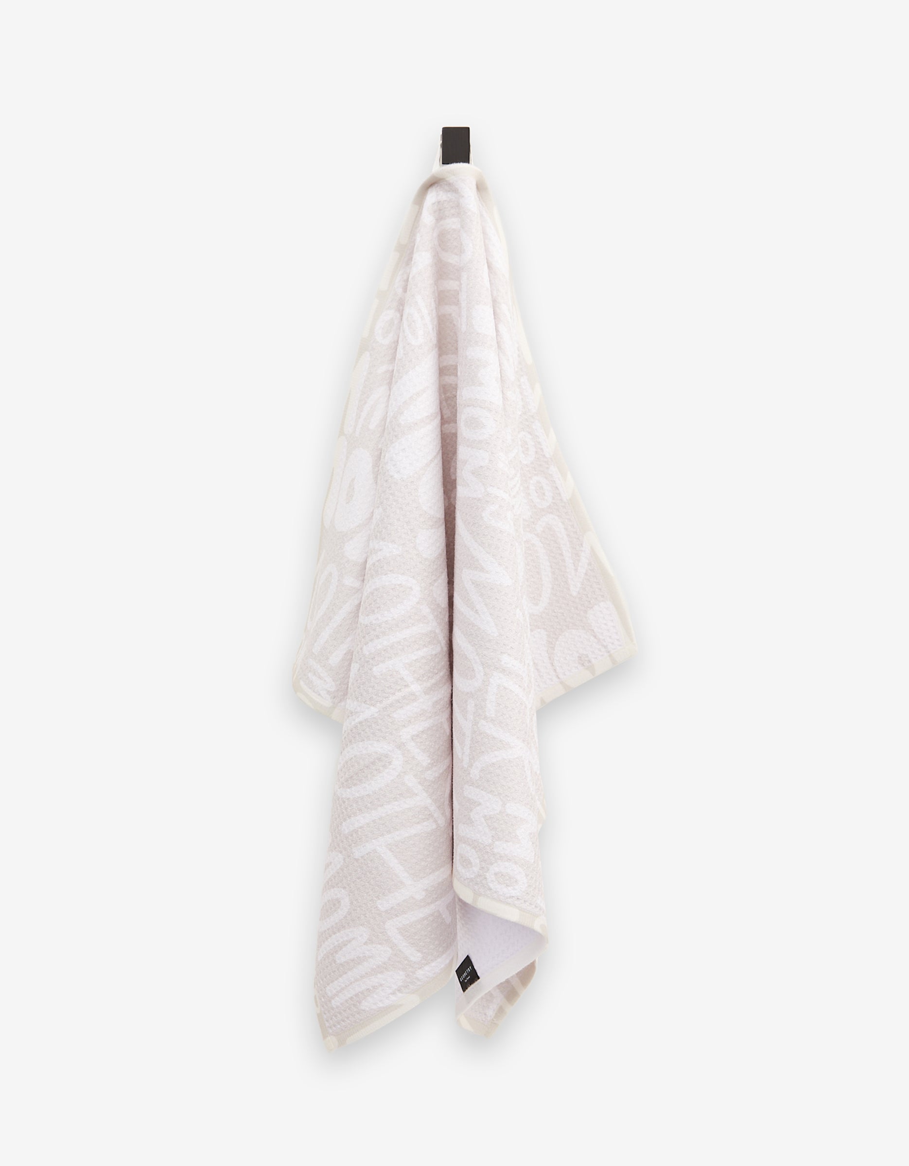 A light-colored, ultra-absorbent GEOMETRY tea towel, called “Mom,” featuring bold white abstract letters and numbers, hangs from a black hook against a plain background.