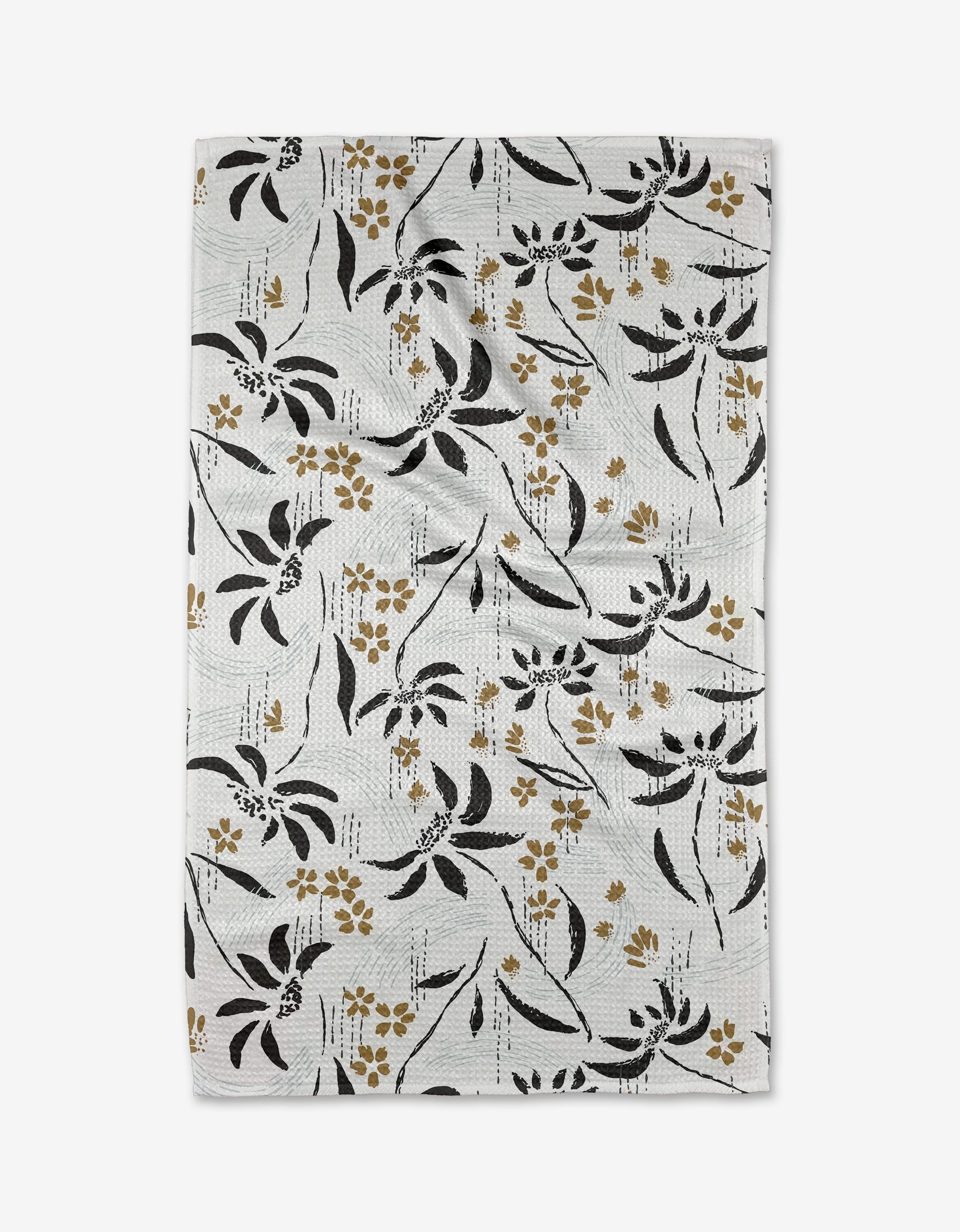The Scenic tea towel by Toboggan Avenue features black and gold floral designs with scattered leaves and small flowers on white Geoweave™ fabric, set against a subtle striped backdrop.