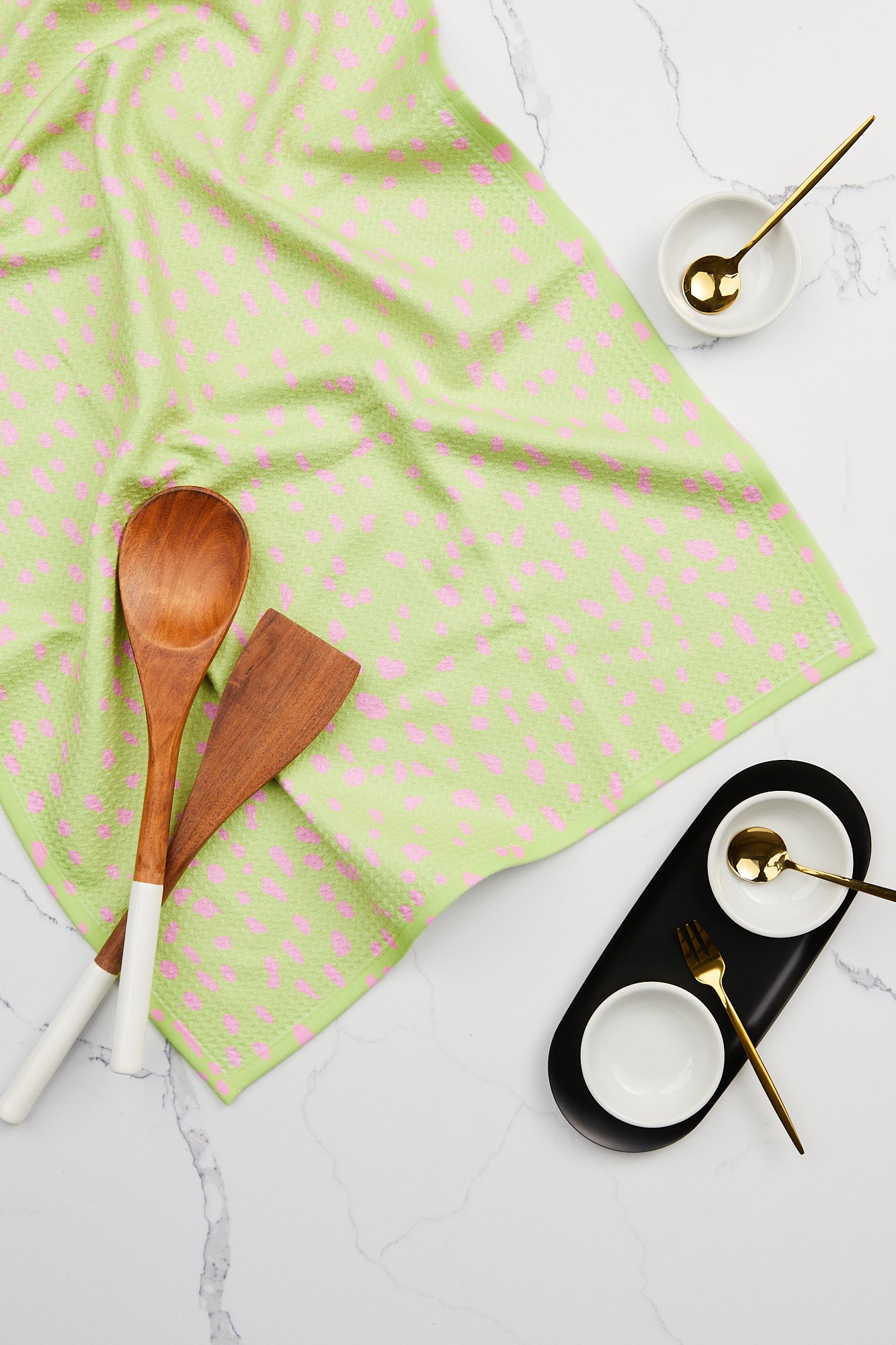 The Speckle Green tea towel by GEOMETRY, made from soft, durable Geoweave fabric, is paired with two wooden utensils with white handles and three small white bowls with gold spoons on a white marble surface.