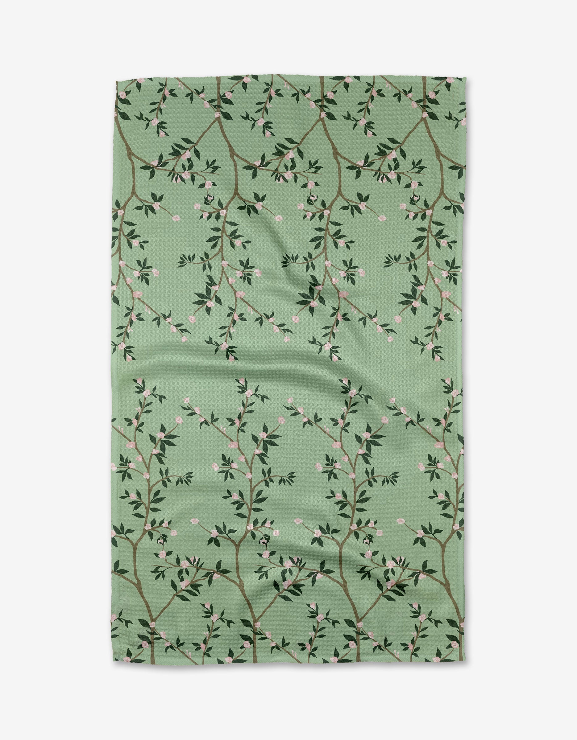 The Lily Tea Towel by Becki Owens features a green Geoweave™ fabric with branches, small leaves, and tiny pink flowers in a lattice pattern, shown slightly wrinkled against a white background.