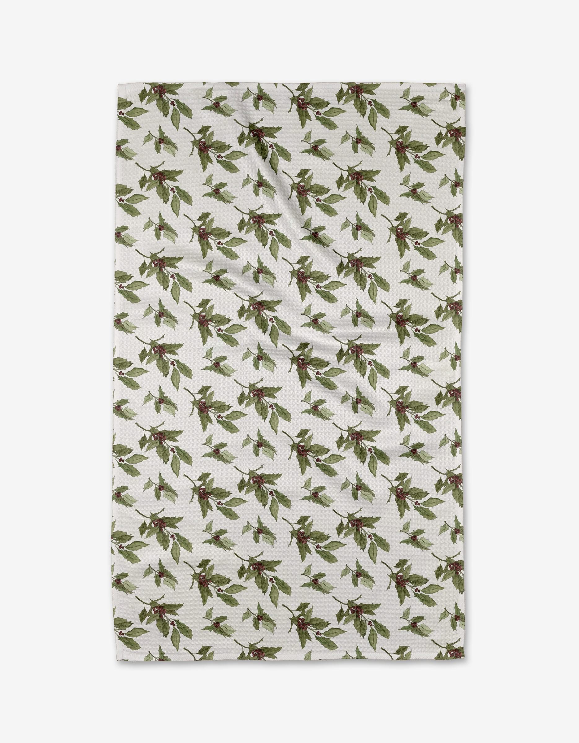 The Aubrey Fairchild Winter Berry kitchen towel is white, made from recycled material, and features a pattern of green leaves and red berries. The towel is slightly wrinkled and shown against a plain white background.