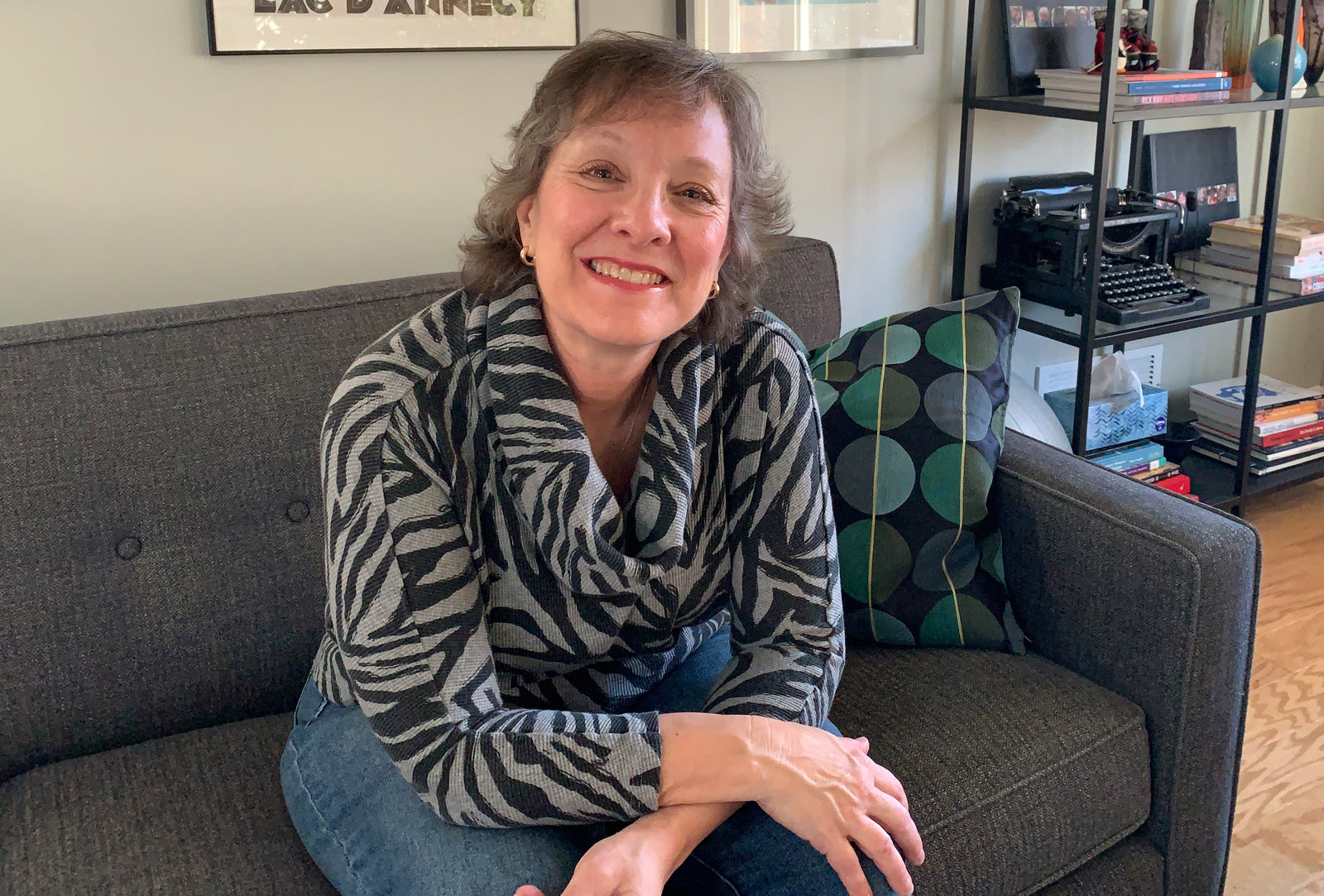A woman with short brown hair, wearing a zebra-print sweater and jeans, sits smiling on a gray couch next to a patterned pillow in a cozy, modern living room.