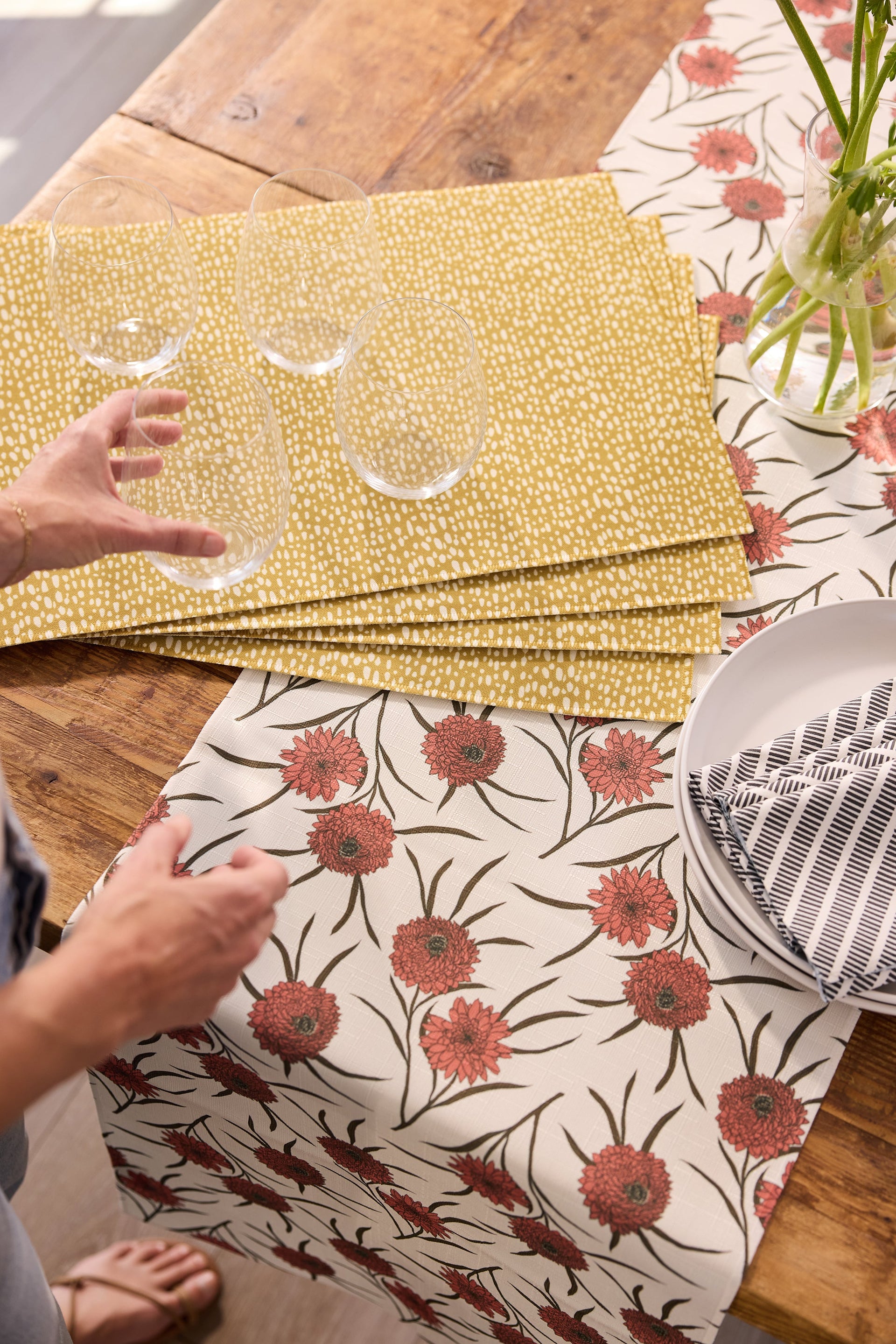 A person sets clear glasses on a stack of yellow placemats atop a wooden table with a cream and red floral table runner, next to a white plate and a vase with flowers.
