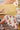 A person sets clear glasses on a stack of yellow placemats atop a wooden table with a cream and red floral table runner, next to a white plate and a vase with flowers.