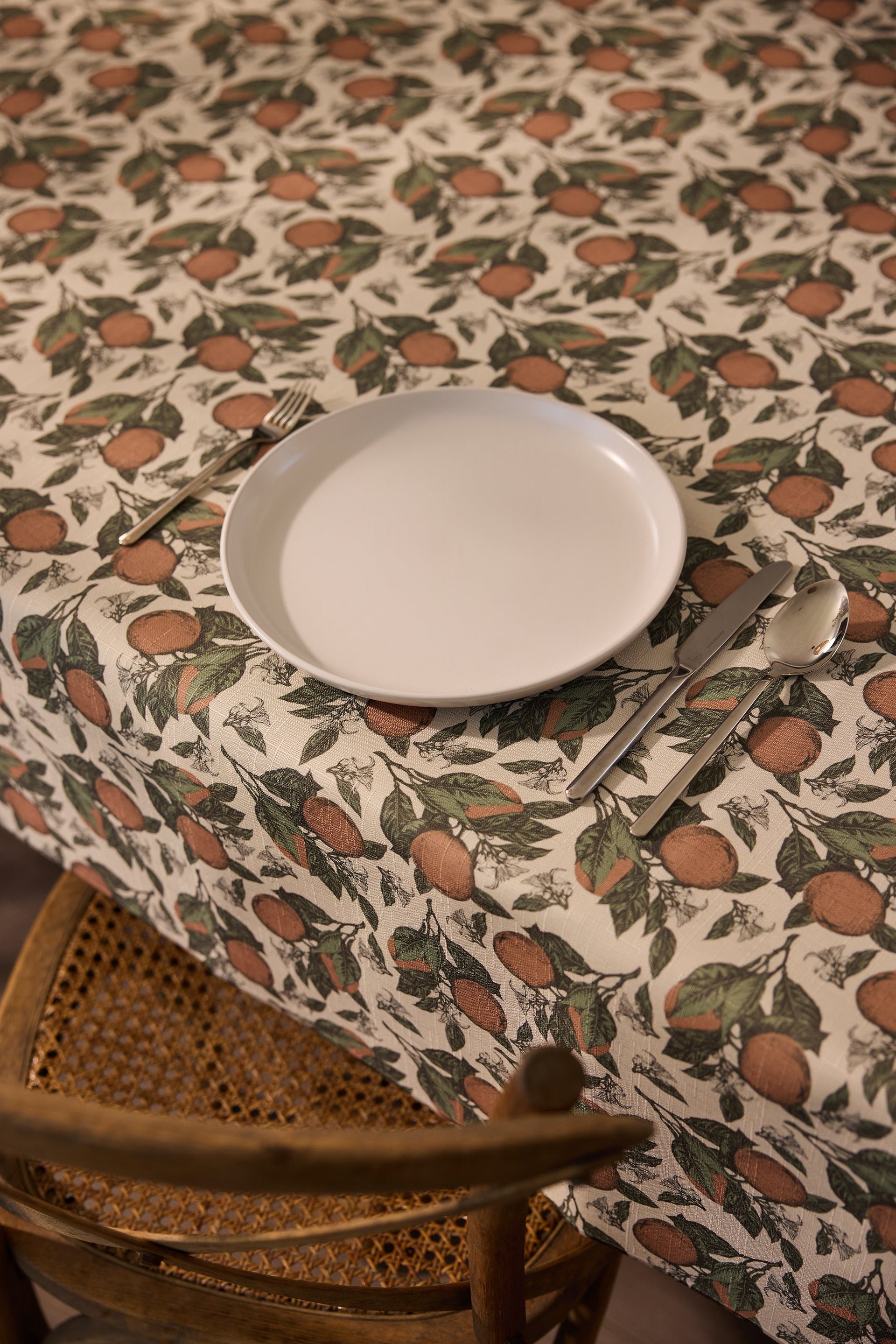 A white plate, fork, knife, and spoon are set on a table with a patterned tablecloth featuring oranges and green leaves; a wooden chair is placed beside the table.