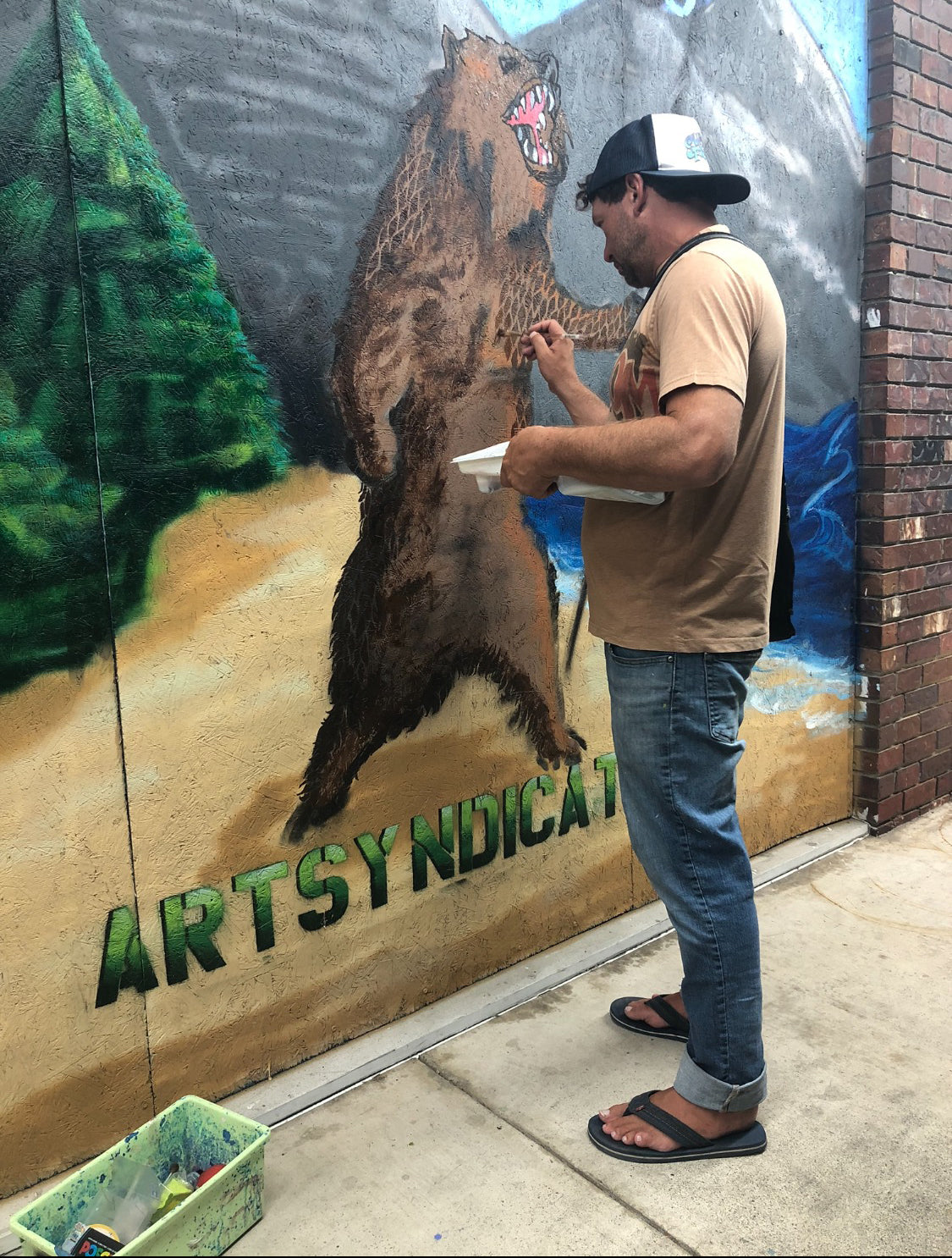 A man in a cap and flip-flops paints a mural of a roaring bear on an outdoor wall. The mural reads ARTSYNDICATE at the bottom, and art supplies are in a tray on the sidewalk nearby.