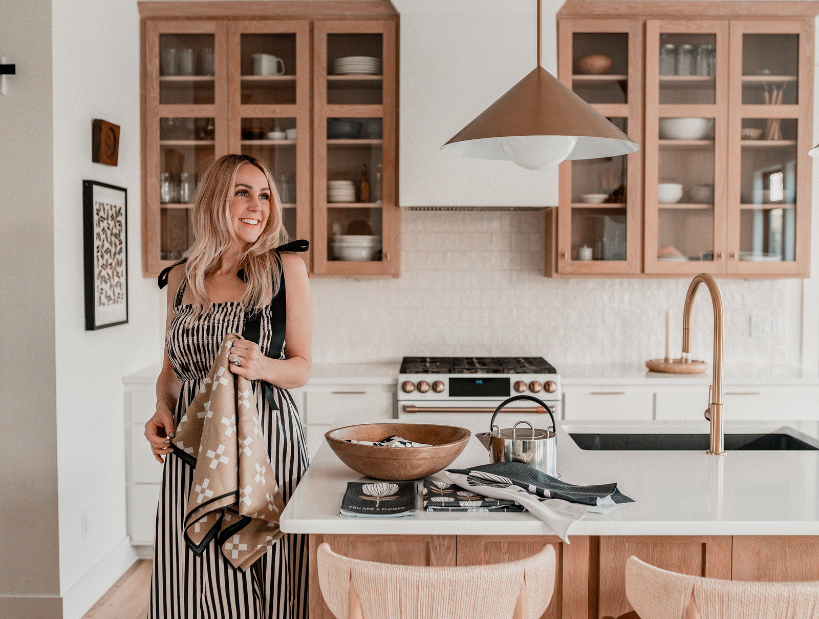 A woman in a striped dress stands smiling in a modern kitchen with wooden cabinets, holding a dish towel beside a kitchen island set with bowls and towels.
