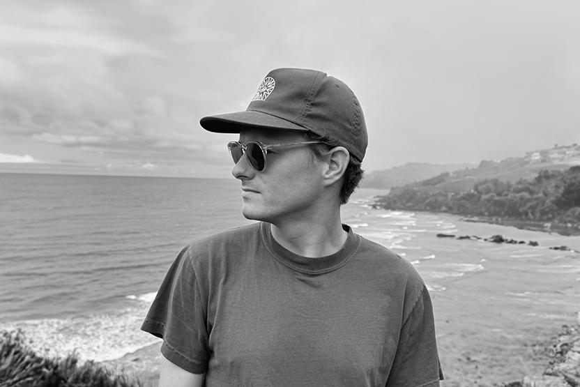 A person wearing sunglasses, a cap, and a t-shirt stands outdoors near a coastline, looking to the side. The scene is in black and white with ocean waves and a cloudy sky in the background.