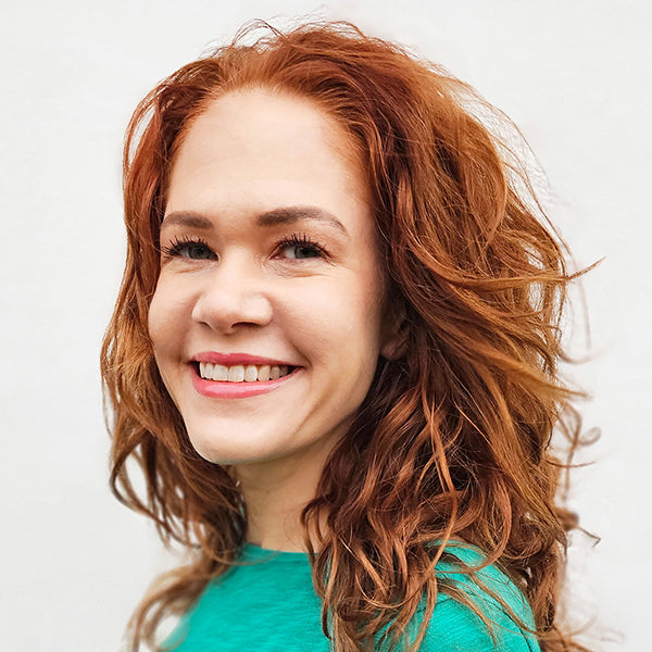 A woman with wavy red hair and fair skin smiles while looking slightly to the side. She is wearing a green top and stands in front of a plain, light-colored background.