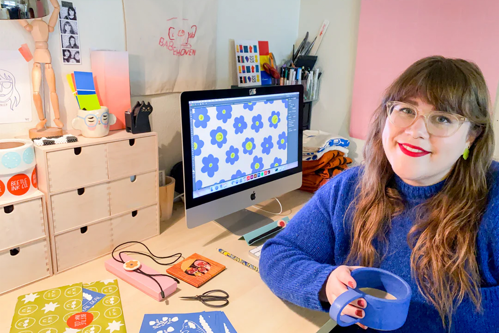 A smiling woman with glasses and a blue sweater sits at a desk holding a blue mug. Behind her is a computer displaying a floral pattern, art supplies, and colorful decorations.