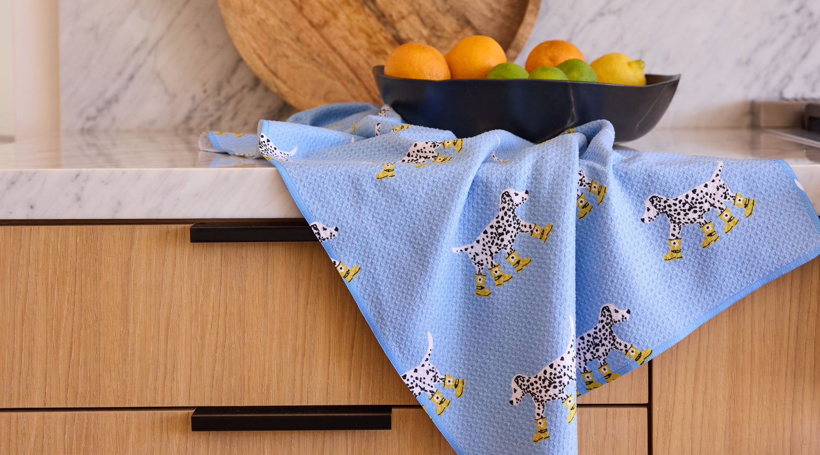 A blue kitchen towel with Dalmatian dog and yellow boot prints draped over a wooden drawer, with a dark bowl of oranges and limes on a marble countertop in the background.
