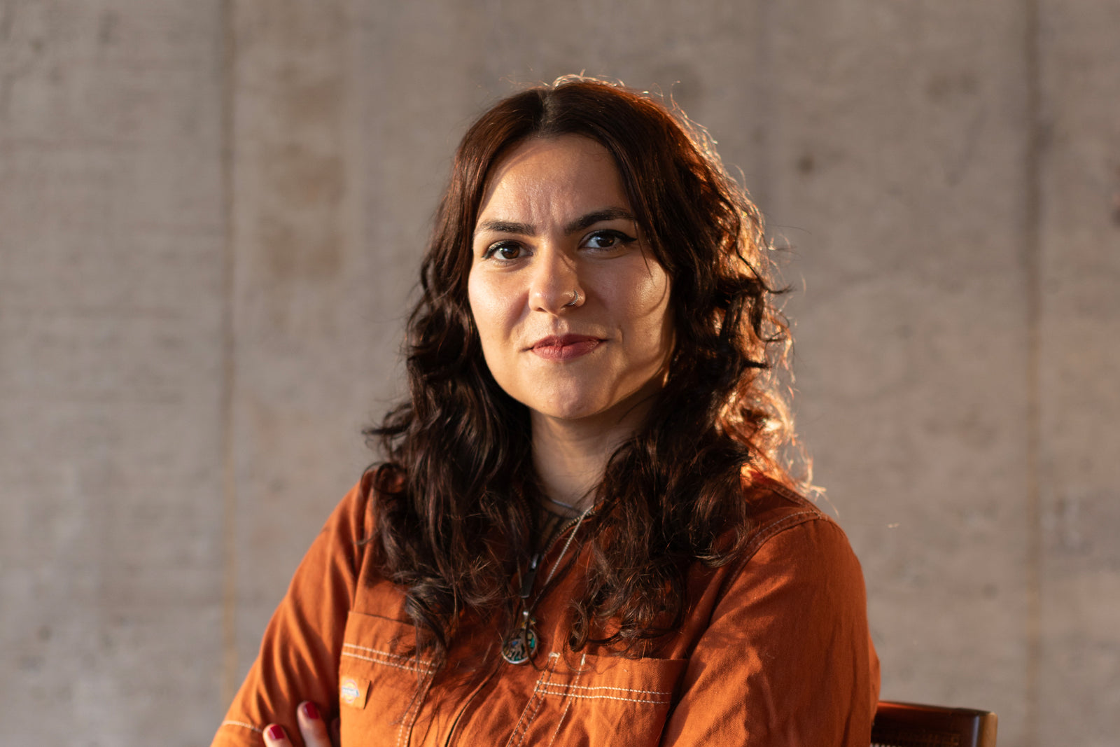 A woman with long curly brown hair wearing an orange shirt sits in front of a plain, light-colored wall, looking confidently at the camera with a slight smile.
