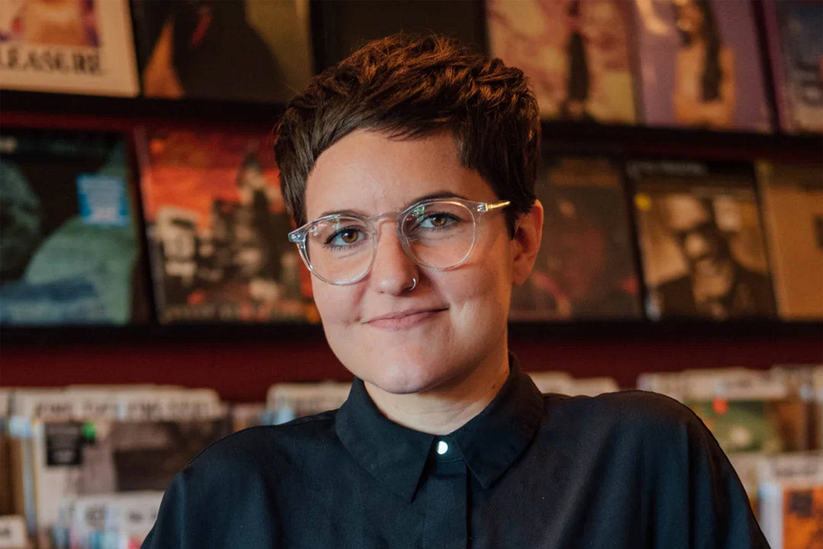A person with short brown hair, clear glasses, and a nose ring smiles slightly while standing in front of shelves filled with vinyl records.