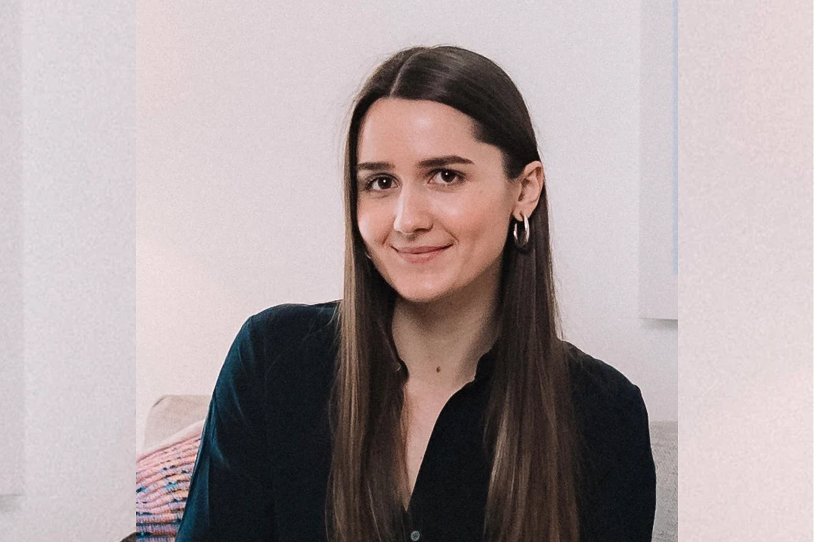 A woman with long straight brown hair, wearing a black top and hoop earrings, smiles gently while sitting indoors against a light-colored background.