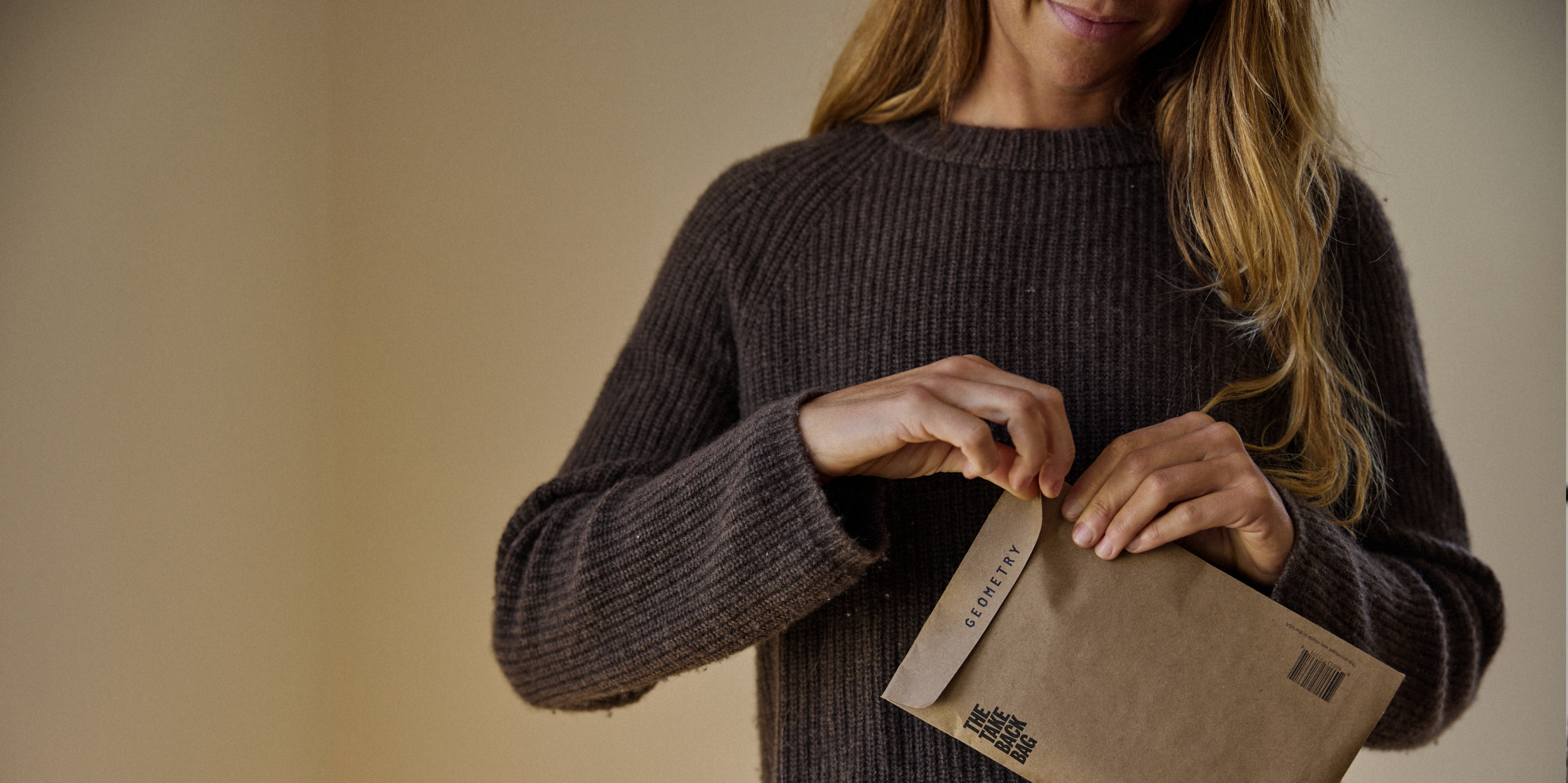 A person with long hair wearing a brown sweater smiles while opening a brown envelope labeled CONFIDENTIAL in black text.