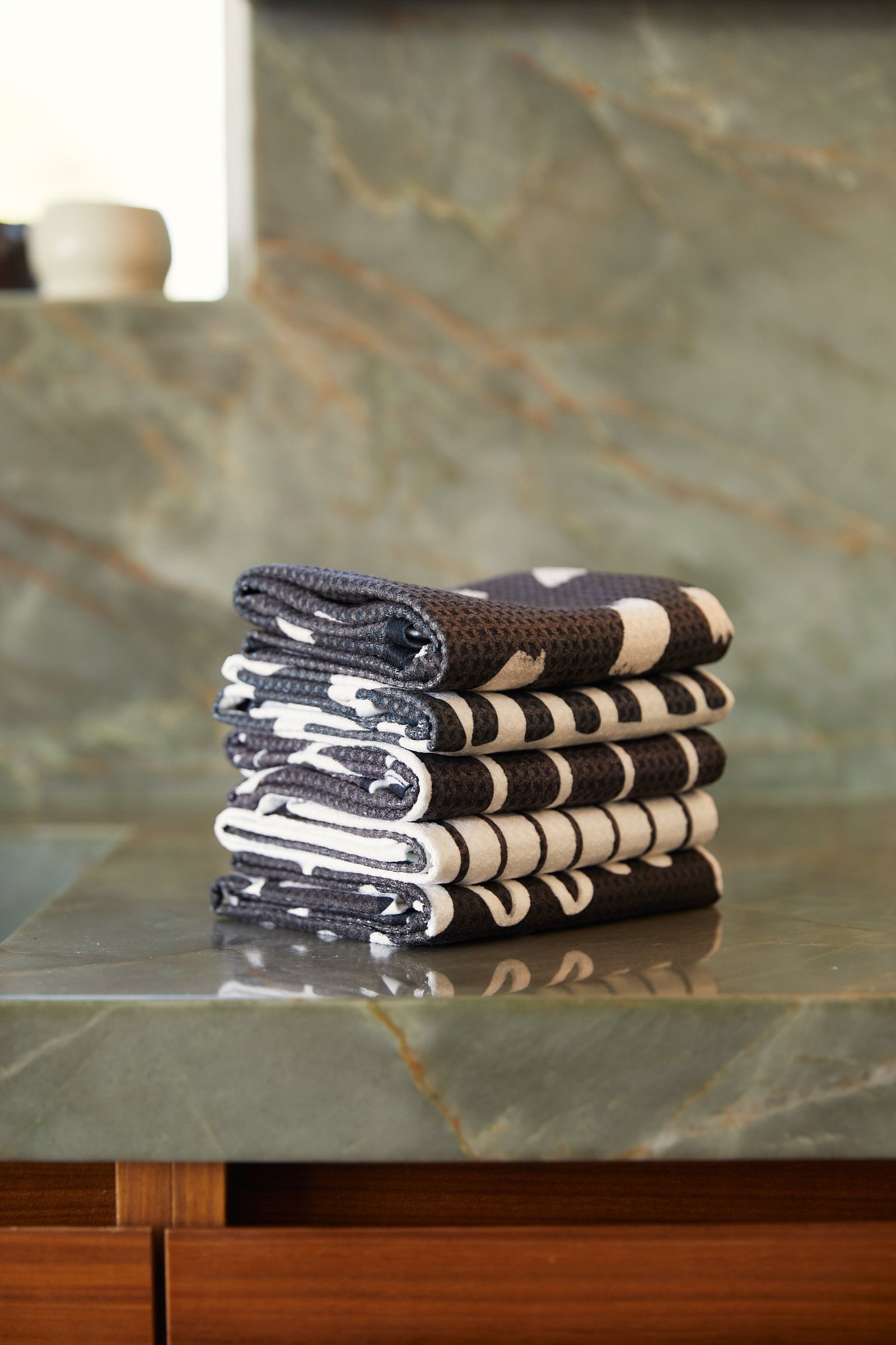 A neatly folded stack of black and white patterned towels sits on a green marble countertop, with a blurred background of a ceramic cup and natural stone surfaces.
