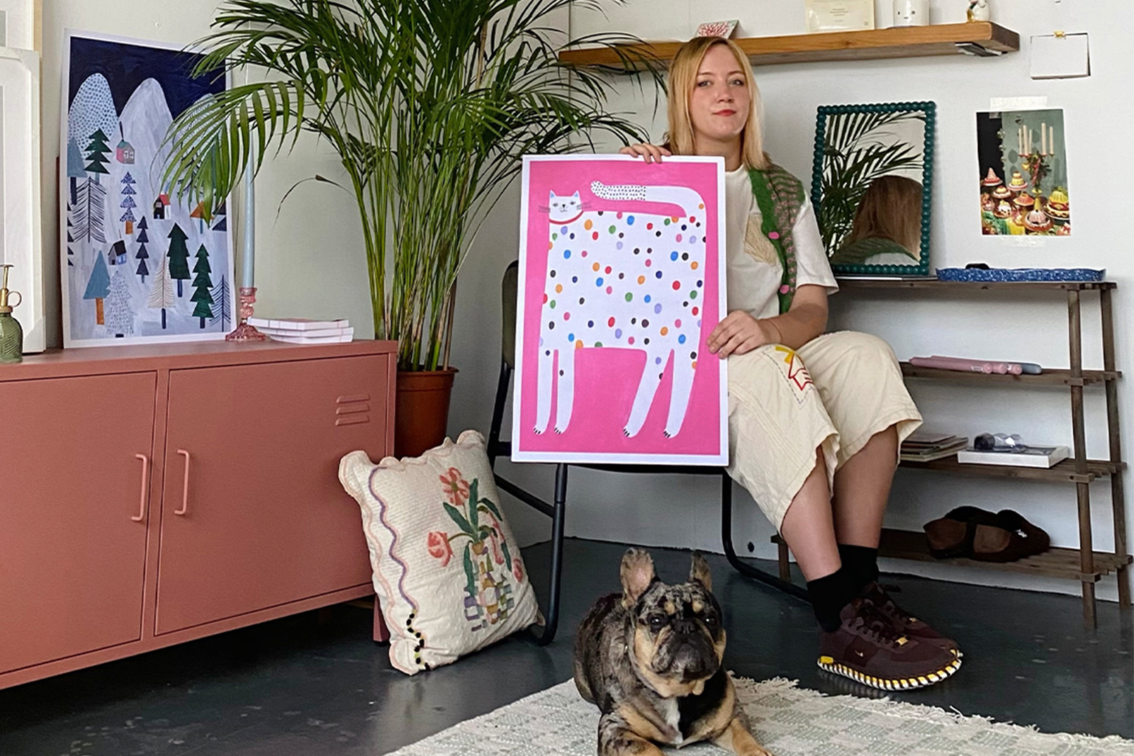A woman sits on a chair holding a colorful painting of a spotted cat on pink, with a small dog lying on the rug in front of her. The room is decorated with art, plants, and cozy decor.