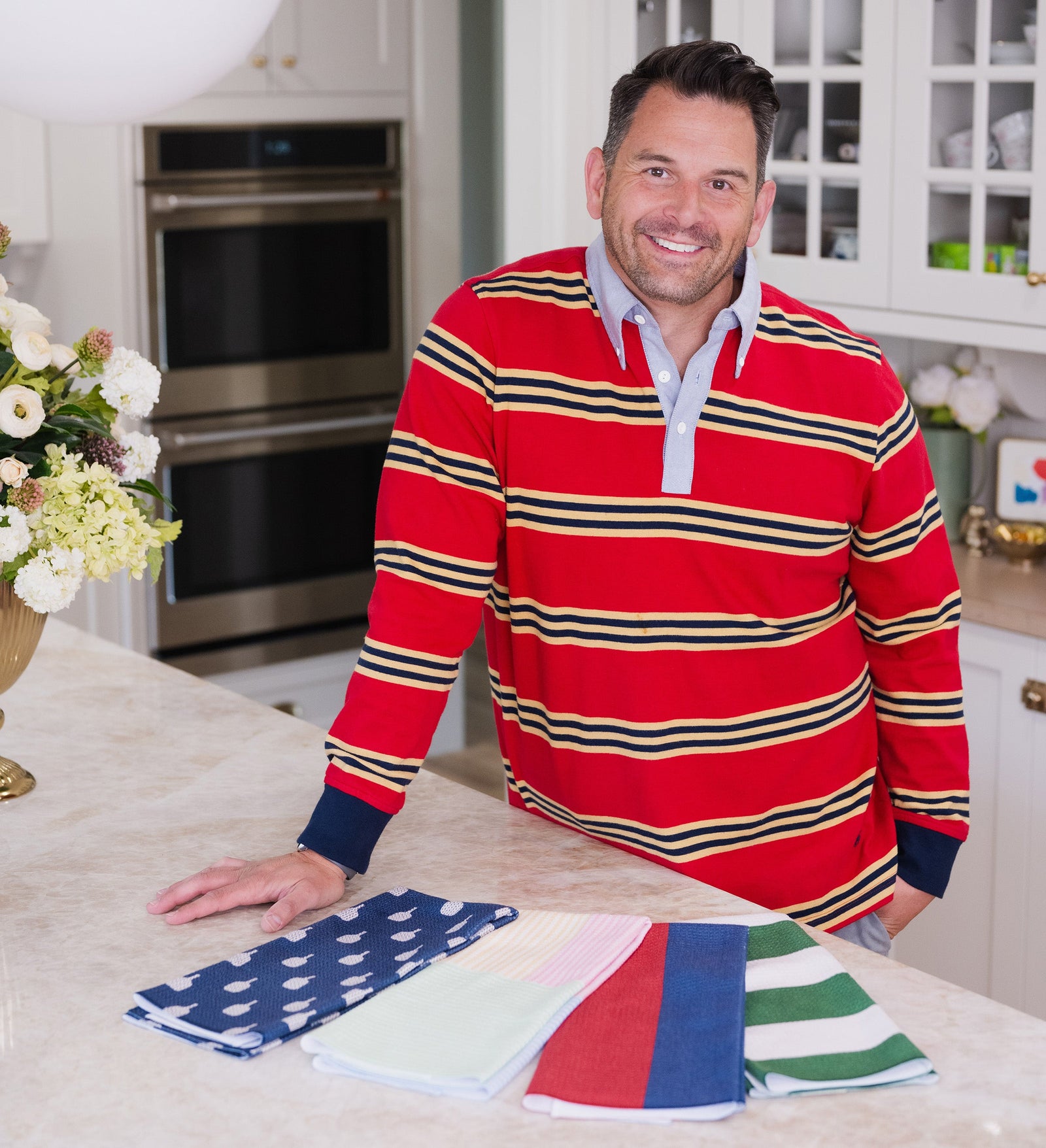A smiling man in a red striped shirt stands in a bright kitchen, leaning on a counter with four neatly folded colorful towels in front of him.