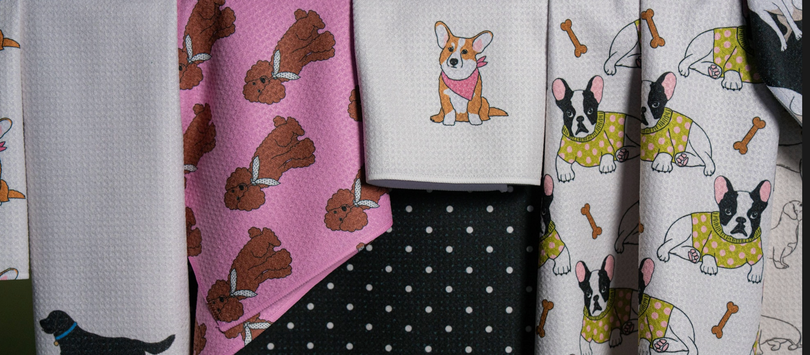 Colorful towels featuring cartoon dogs: brown poodles on pink, a single corgi in a scarf, and French bulldogs with bones on light backgrounds, hanging in front of a black polka dot cloth.
