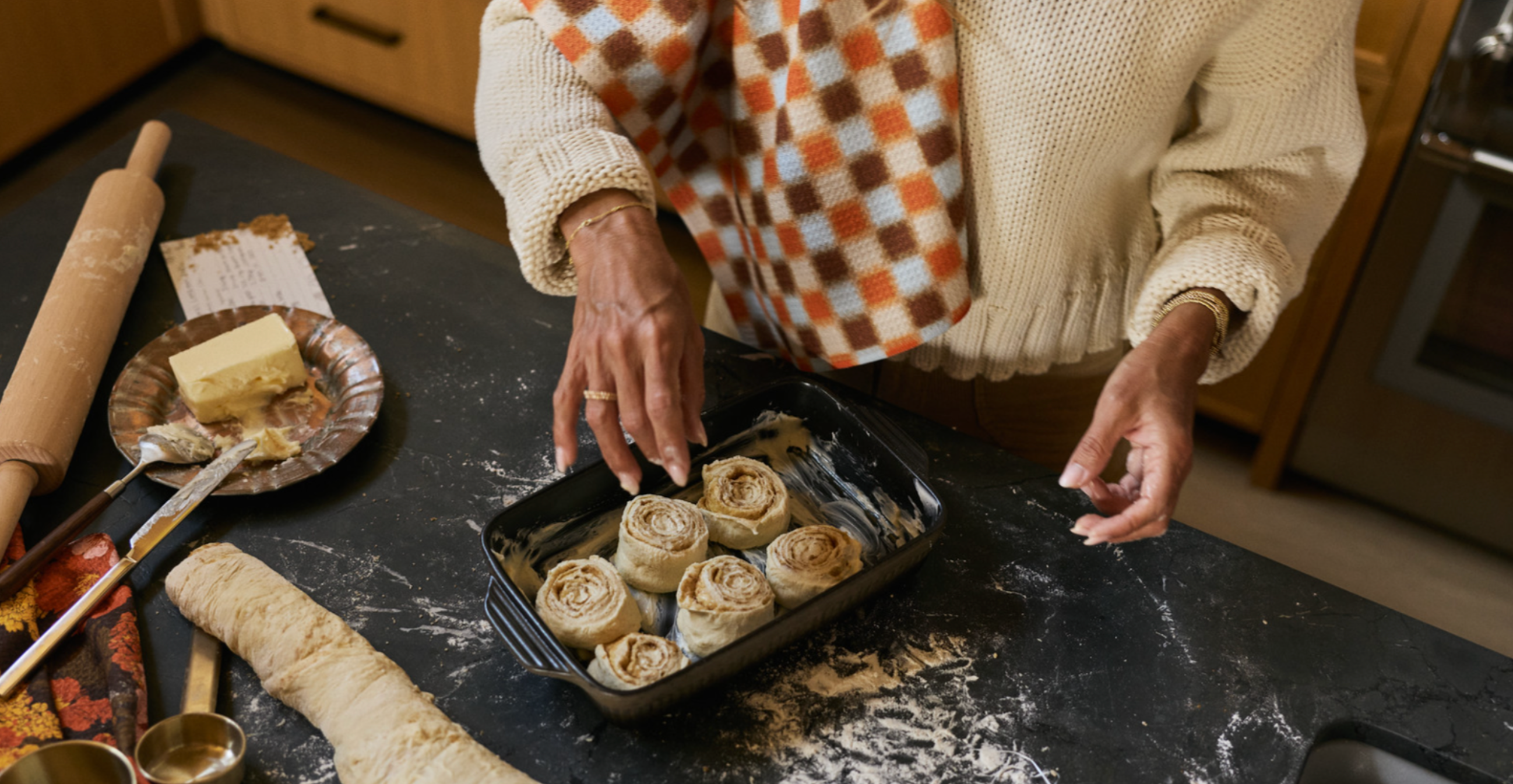 A person wearing a striped apron and jeans tucks a pumpkin-patterned dish towel into the apron, standing in a kitchen with wooden cabinets in the background.