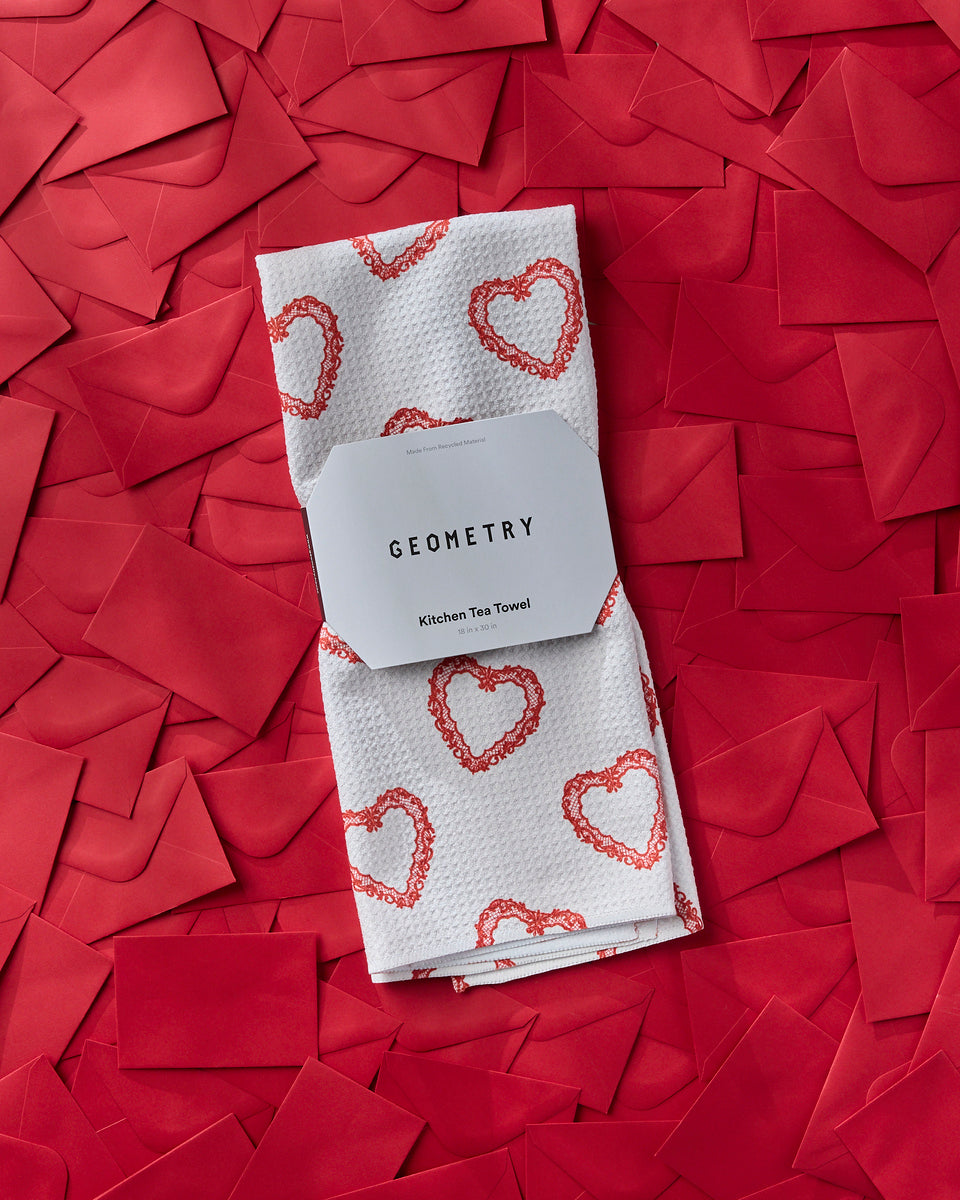 A red heart-shaped box of assorted chocolates sits beside a red envelope on a pink and white checkered cloth, on a light gray surface.