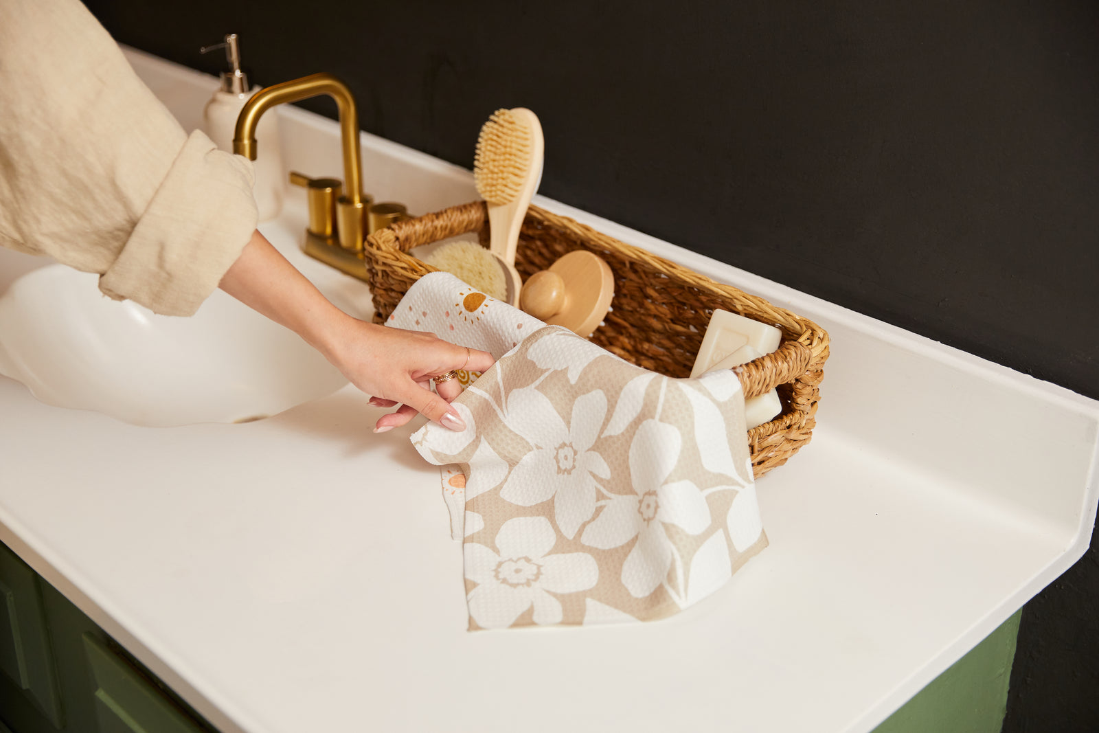 A hand reaches for a patterned cloth near a wicker basket holding toiletries, such as a brush and lotion, on a white bathroom countertop with a gold faucet.