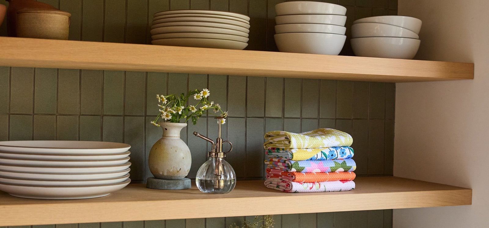 Two wooden shelves hold stacked white dishes, bowls, a vase with small flowers, a glass mister, and a neatly folded stack of colorful patterned dish towels against a green tiled wall.