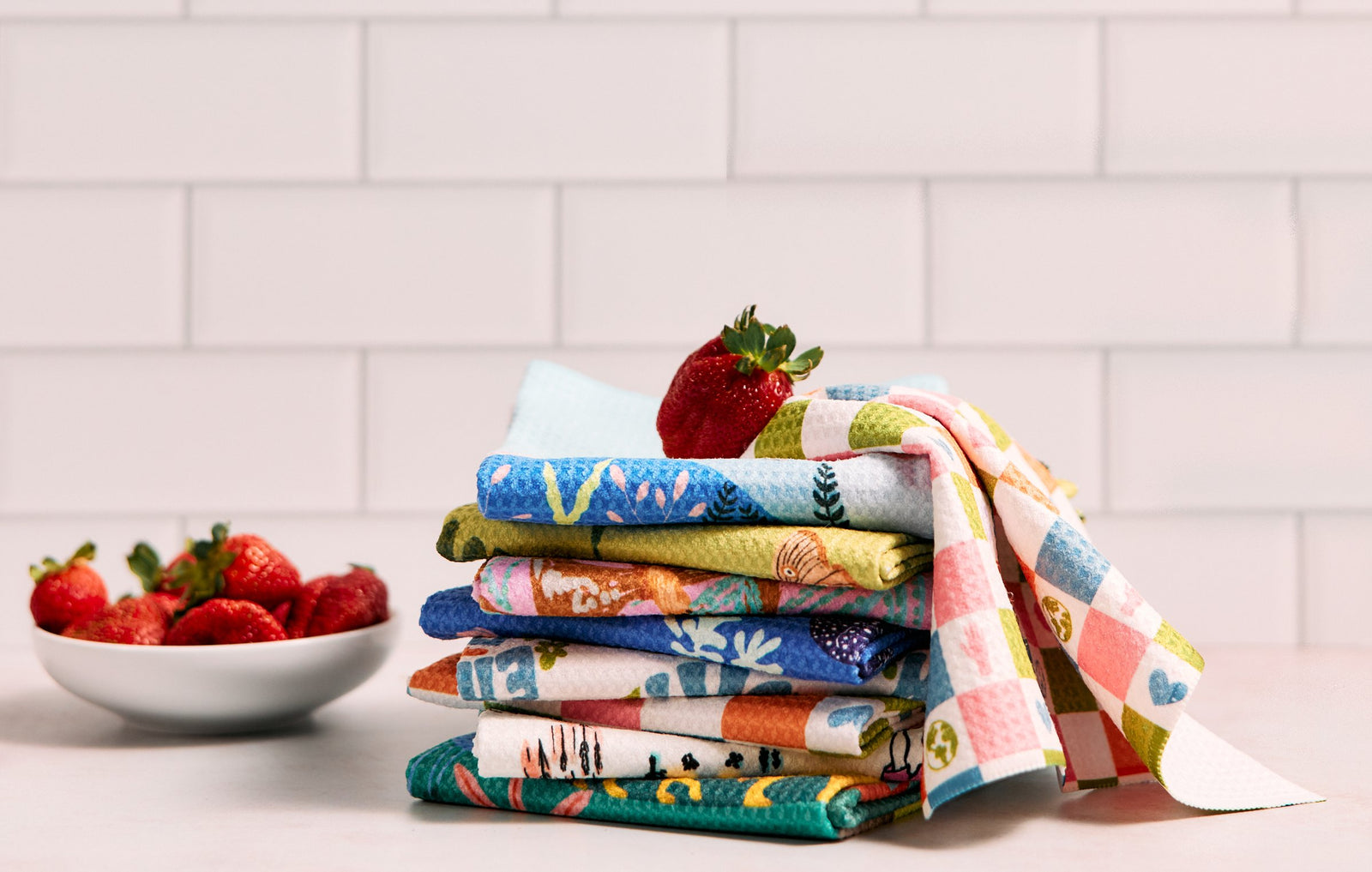 A stack of colorful, patterned dish towels with a strawberry on top sits on a counter, next to a white bowl filled with strawberries, against a white tiled background.