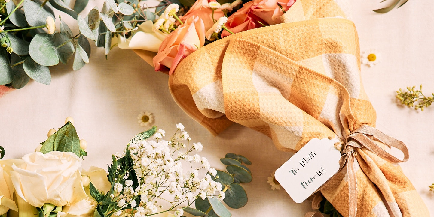 A bouquet of pink and white roses wrapped in a yellow checkered cloth with a tag reading To: Mom, From: Us, surrounded by eucalyptus leaves and baby’s breath flowers on a light surface.