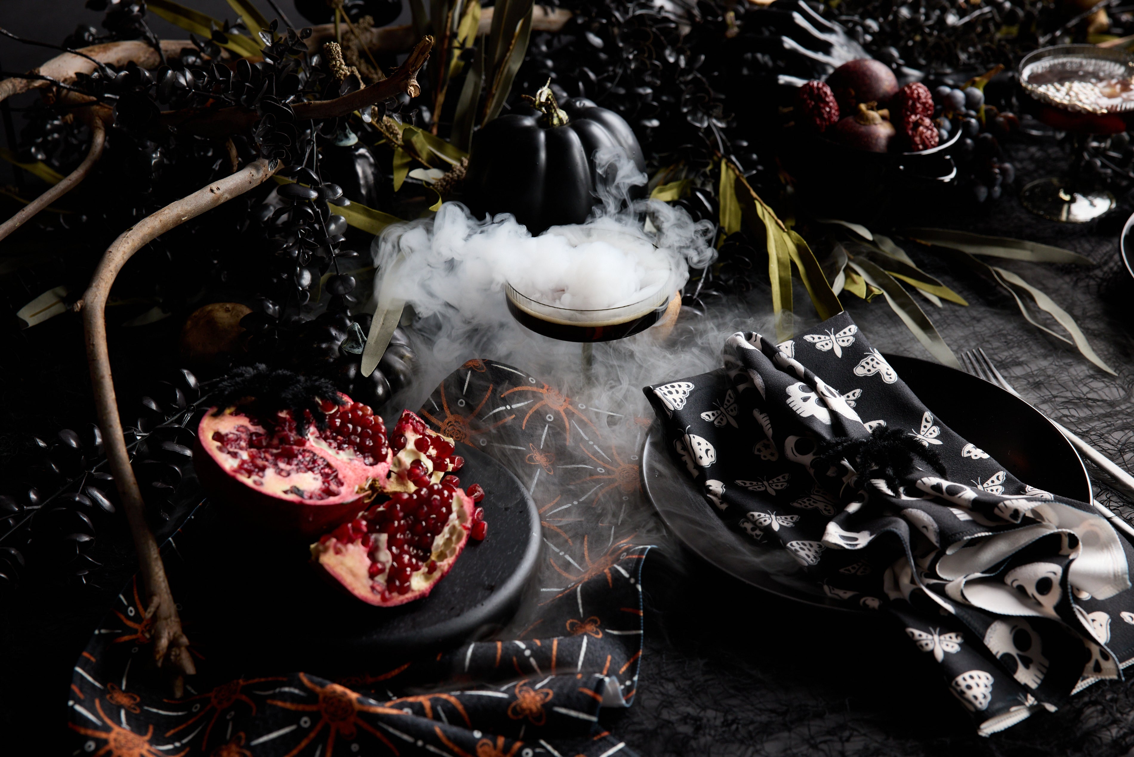 A dark, moody Halloween-themed table with a halved pomegranate, smoky cocktail, black dishes, and napkins decorated with skull and insect patterns, surrounded by dramatic black foliage.