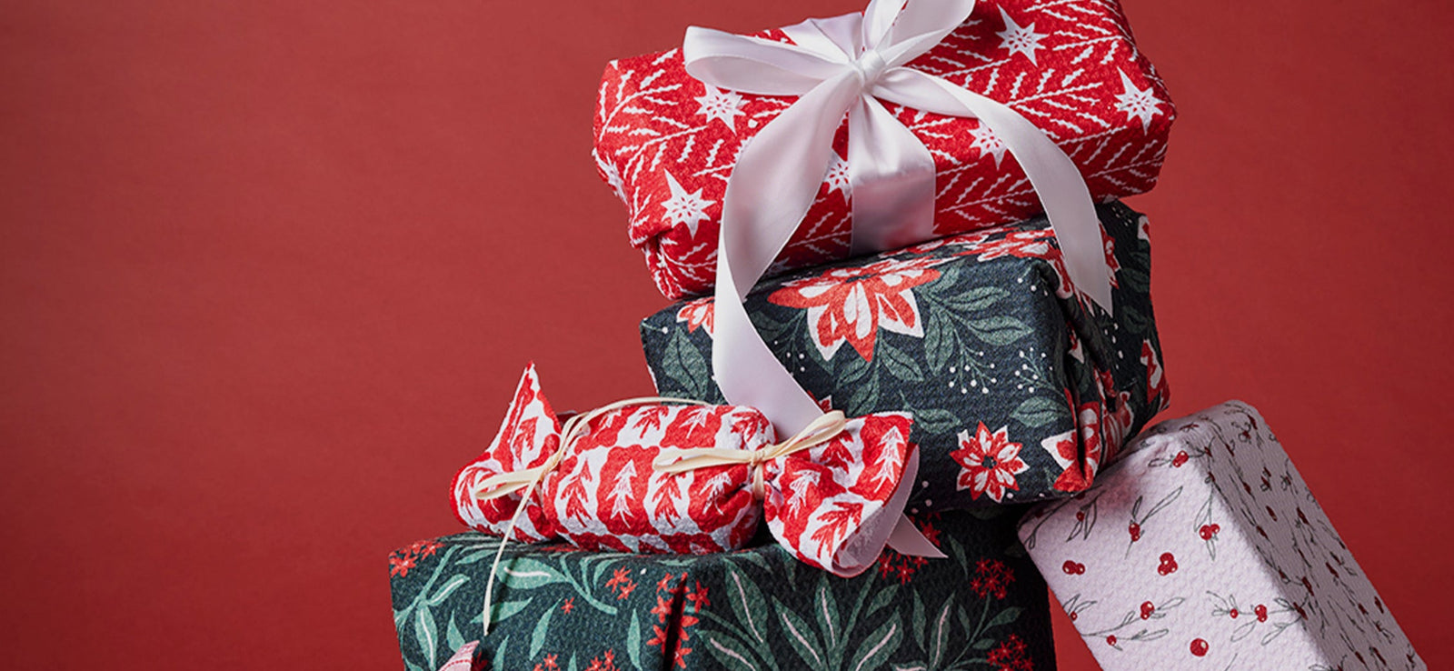 A stack of five wrapped gifts in festive, patterned paper with red, green, and white designs, topped with a white ribbon bow, set against a plain red background.
