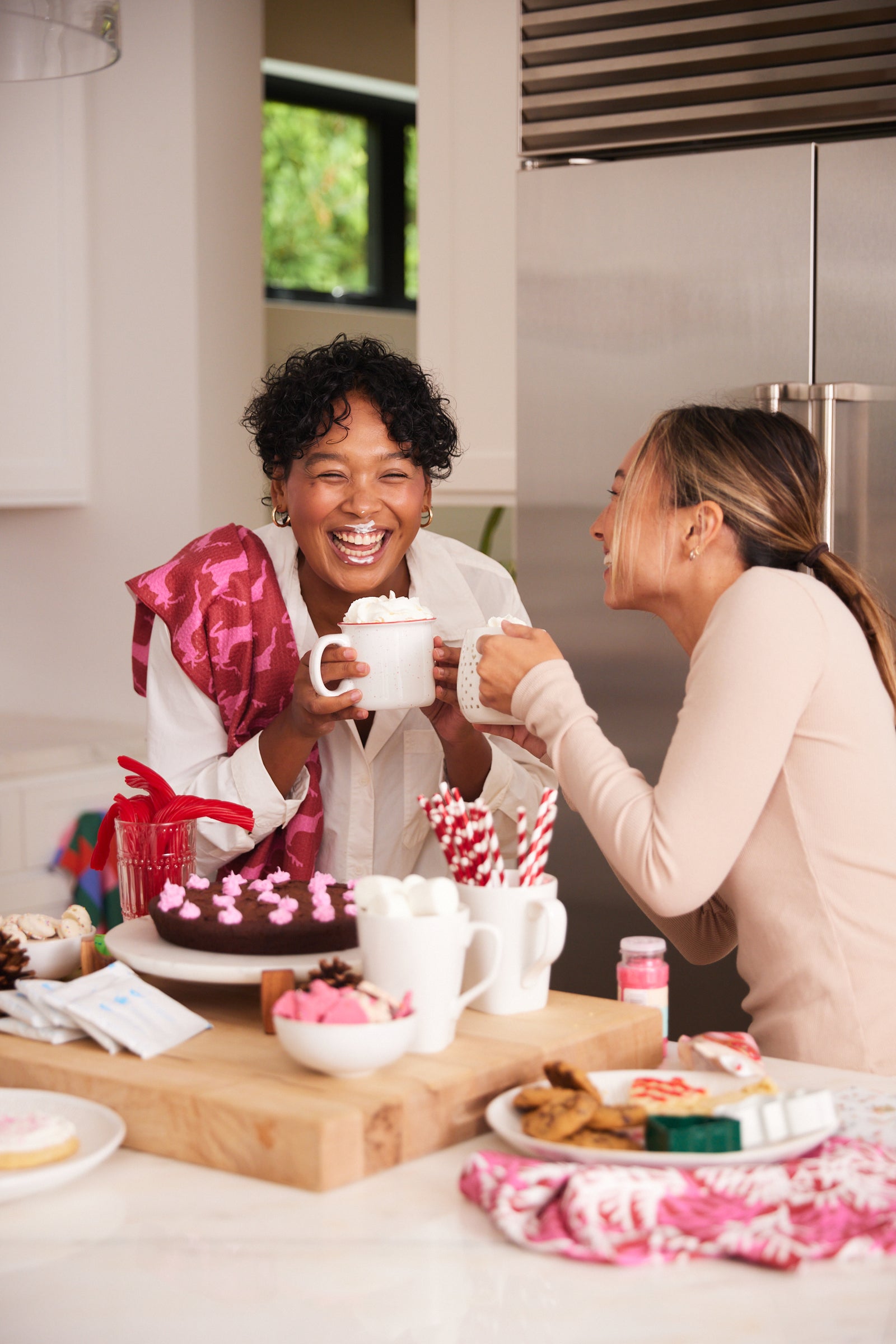 Two women laugh and enjoy mugs of hot chocolate in a bright kitchen, surrounded by sweet treats, desserts, and festive decorations on the counter.