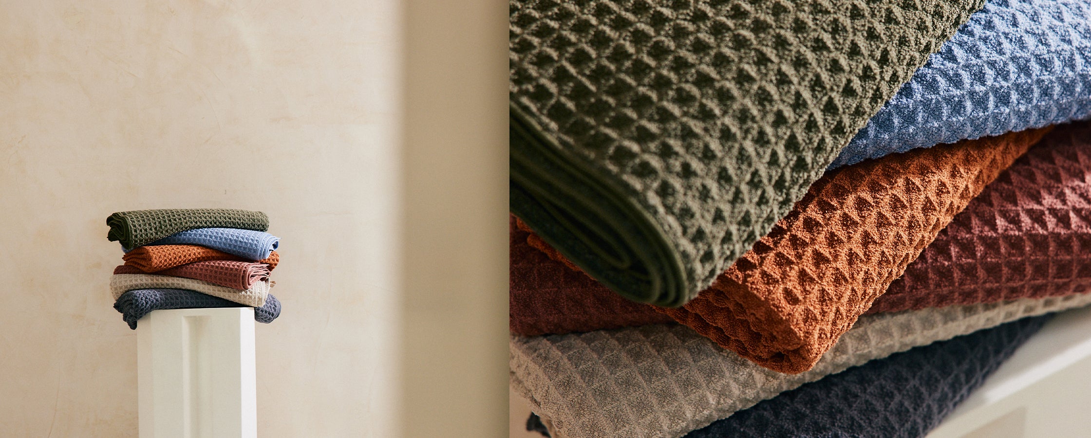 A stack of waffle-textured towels in various colors, including green, blue, rust, and beige, is neatly arranged on a shelf. A small bundle of rolled towels sits on a white surface nearby.