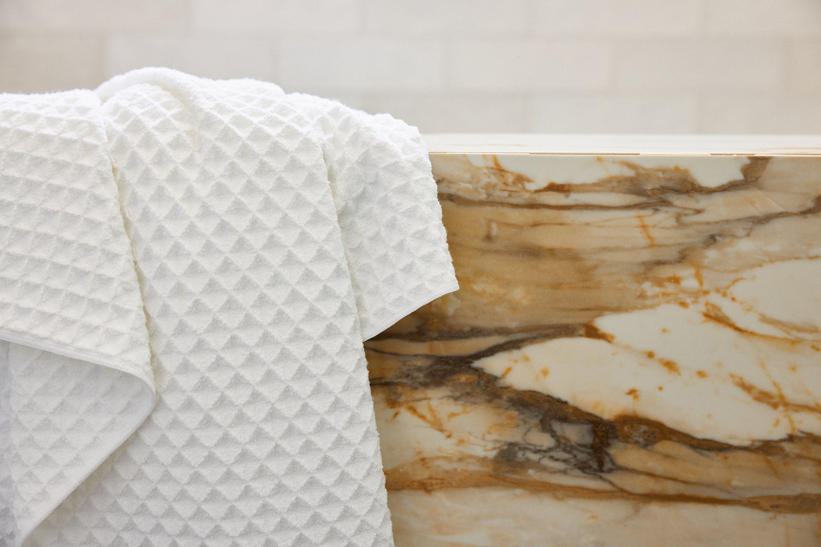 A white textured towel hangs over the edge of a marble bathtub with brown and gold veining, set against a light-colored tiled wall.