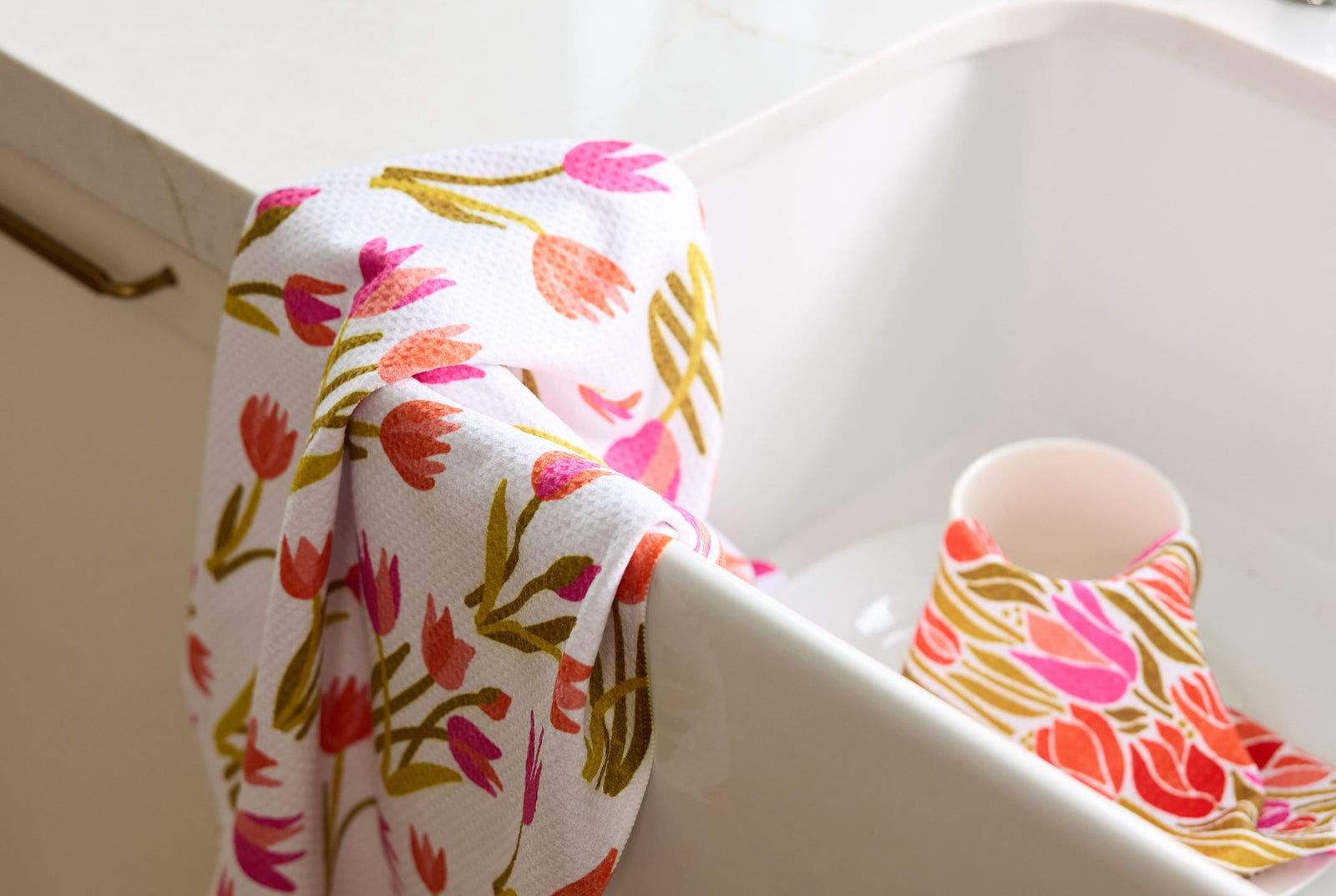 A white dish towel with colorful floral prints drapes over the edge of a white sink. Inside the sink, theres a white mug with a matching floral cloth partially covering it.