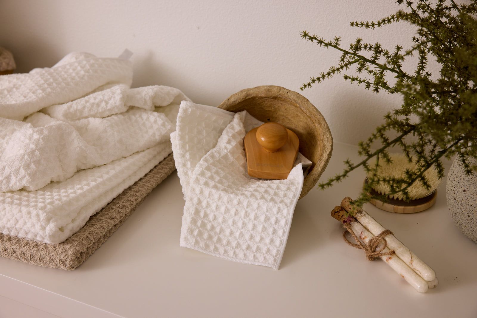 A stack of white textured towels and a folded cloth with a wooden massage tool sit on a white surface, next to a plant and tied candles, creating a calm, spa-like setting.