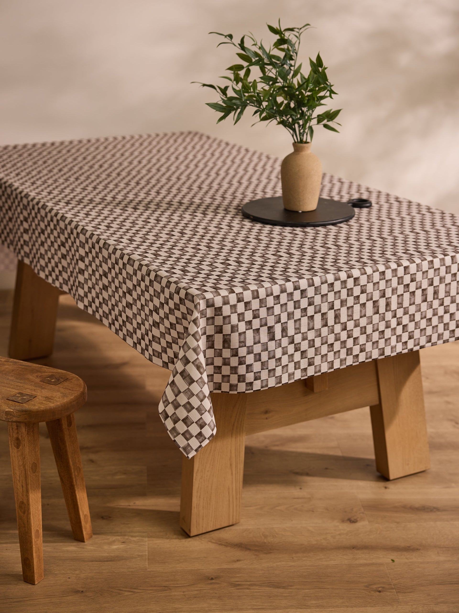A wooden table covered with a checkered tablecloth, a small vase with green leaves on a black tray, and a wooden stool on a light wooden floor.