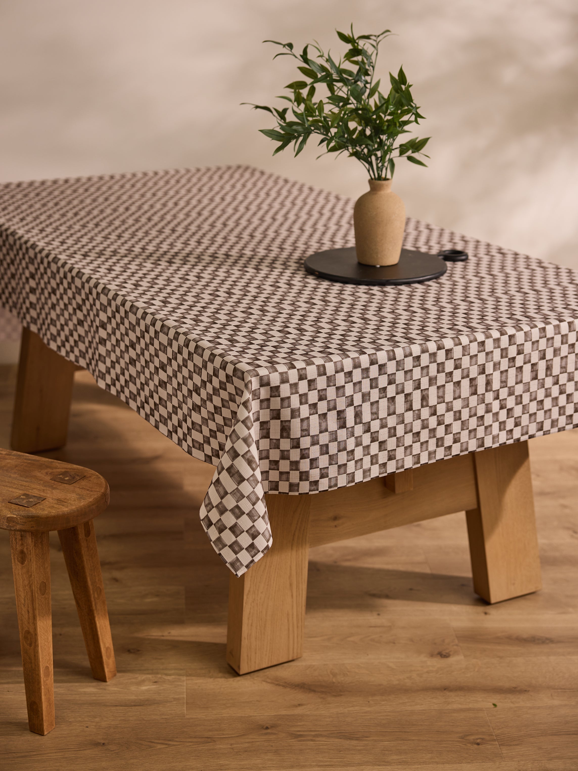 A wooden table covered with a checkered tablecloth, a small vase with green leaves on a black tray, and a wooden stool on a light wooden floor.