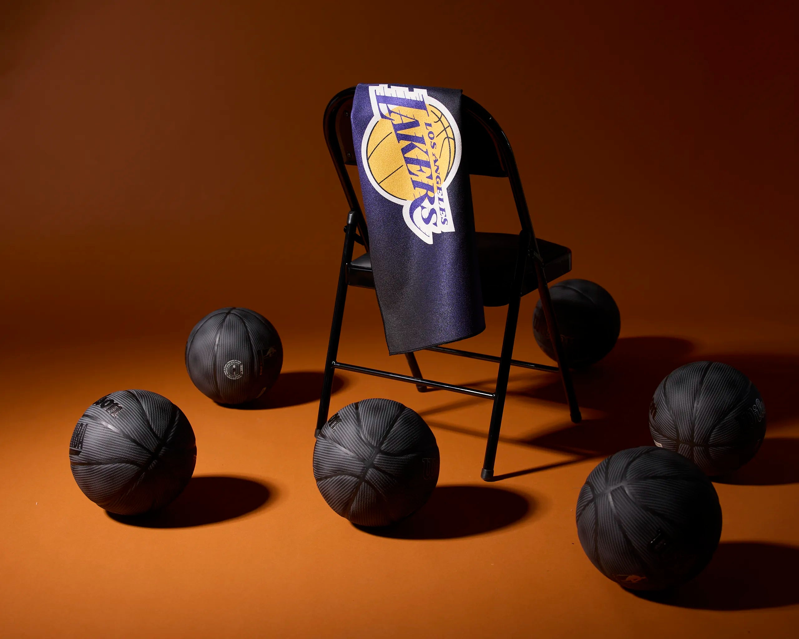 A black folding chair with a Los Angeles Lakers towel draped over the back is surrounded by six black basketballs on an orange-brown background.