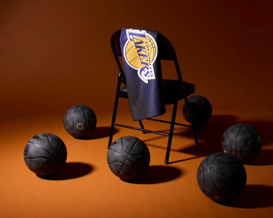 A black folding chair with a Los Angeles Lakers towel draped over the back is surrounded by six black basketballs on an orange-brown background.